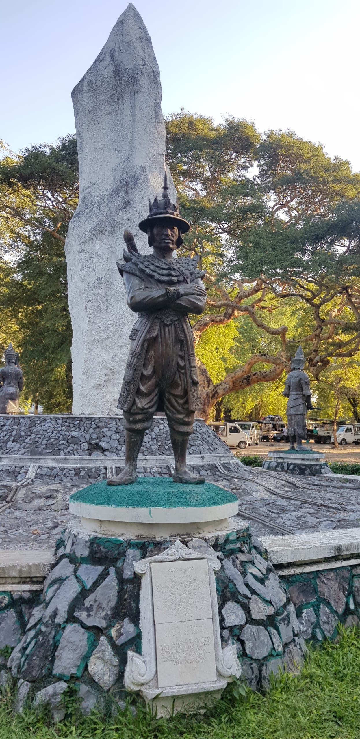 Bronze standing statue of General Maha Bandula in plumed helmet with arms crossed over a sword, on a stone pedestal before a tall stele