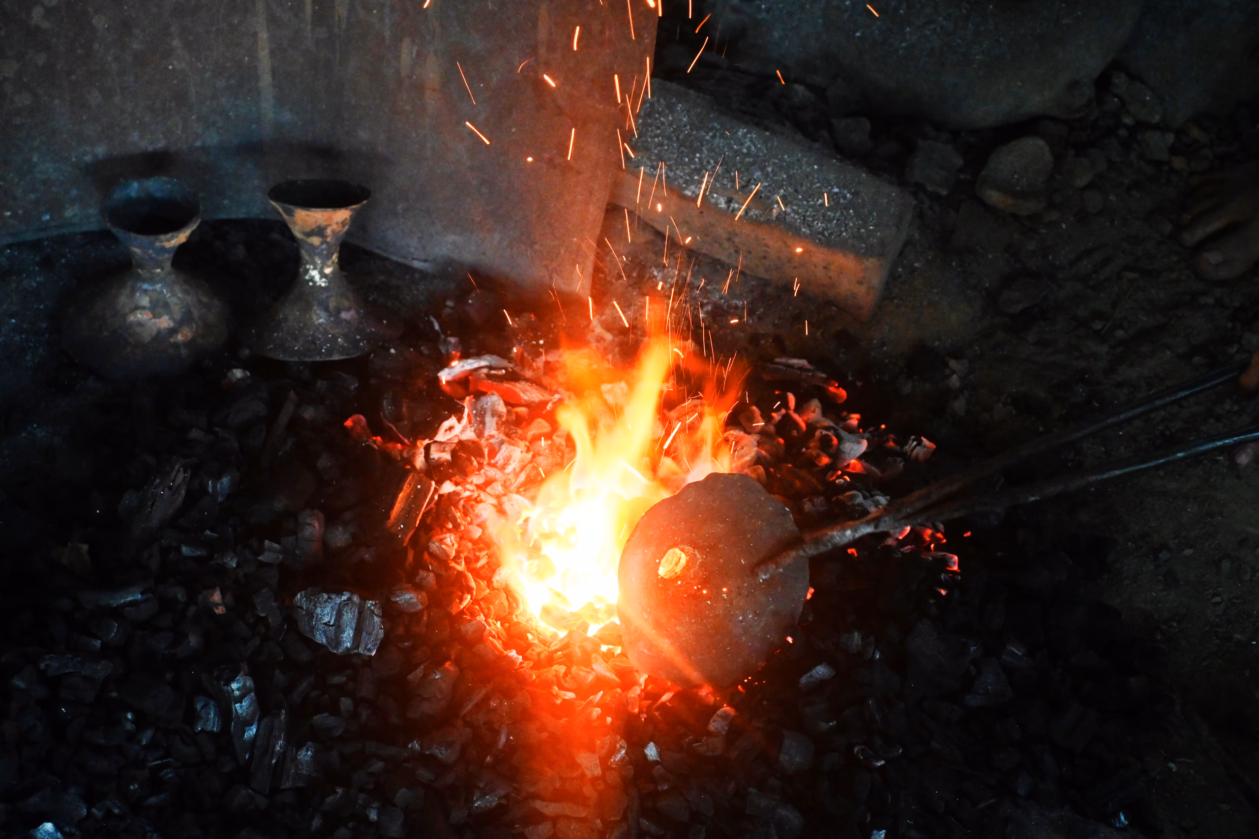 A rounded lump of bell-metal held on an iron rod glows in a bed of burning charcoal, sparks rising; two finished bell-metal vessels sit in the dark at the back of the forge