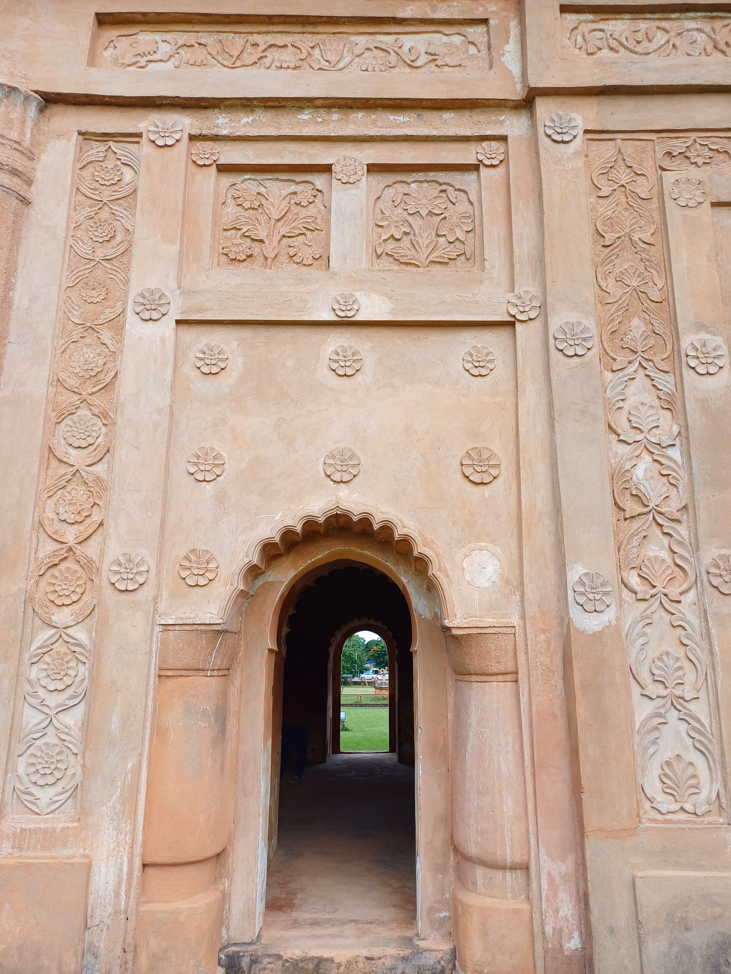 Close view of the Rang Ghar's brick wall at Sivasagar, covered in moulded terracotta ornament: bands of rosettes, framed floral relief panels, and a scalloped arched niche opening to greenery beyond