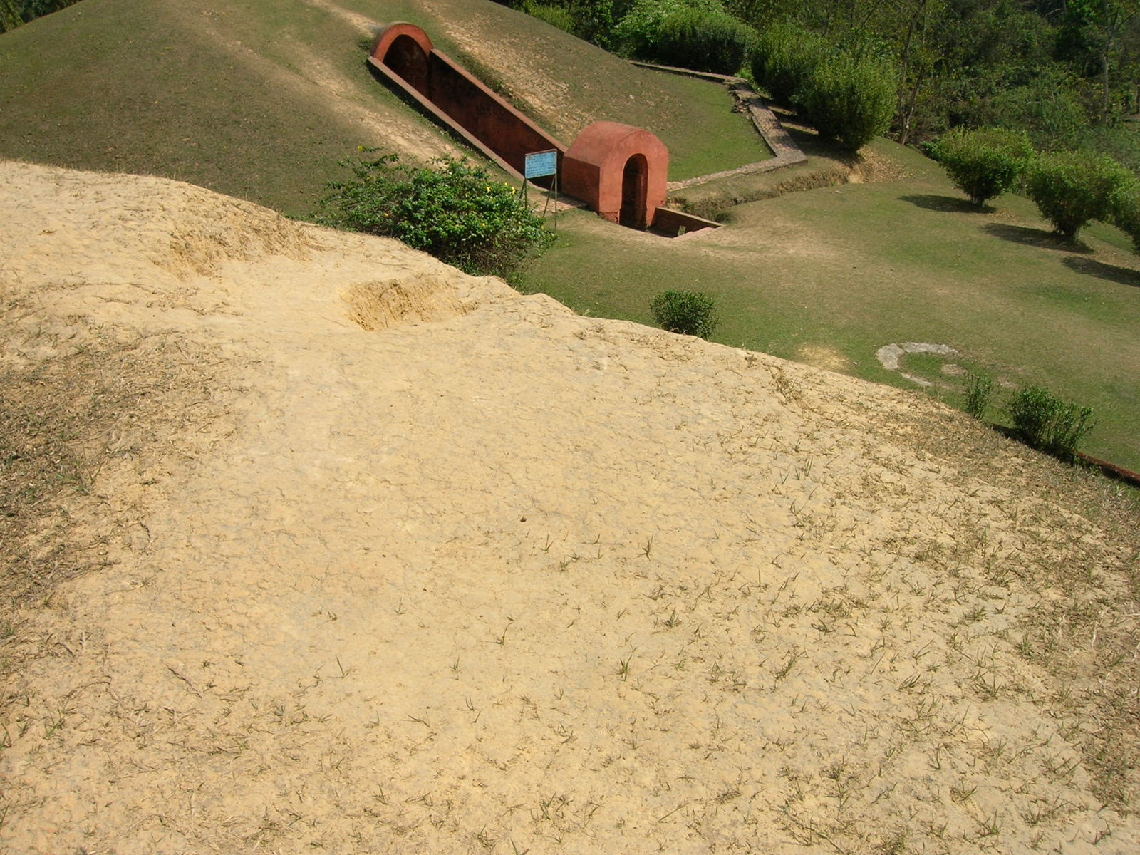 Looking down the grassy crown of one earthen burial mound toward the red brick arched vault entrance of an adjacent maidam, set into a mowed lawn with shrubs and a small signboard