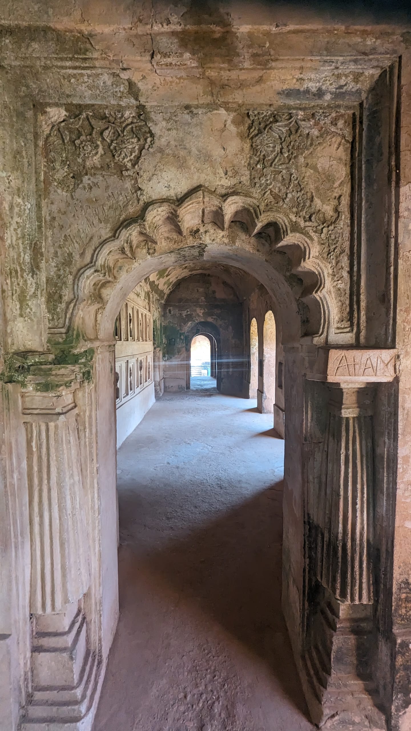 A scalloped multifoil brick arch inside the Talatal Ghar, carved plaster floral spandrels above it and a second arched hall with niched walls visible beyond, leading to a small lit opening at the far end