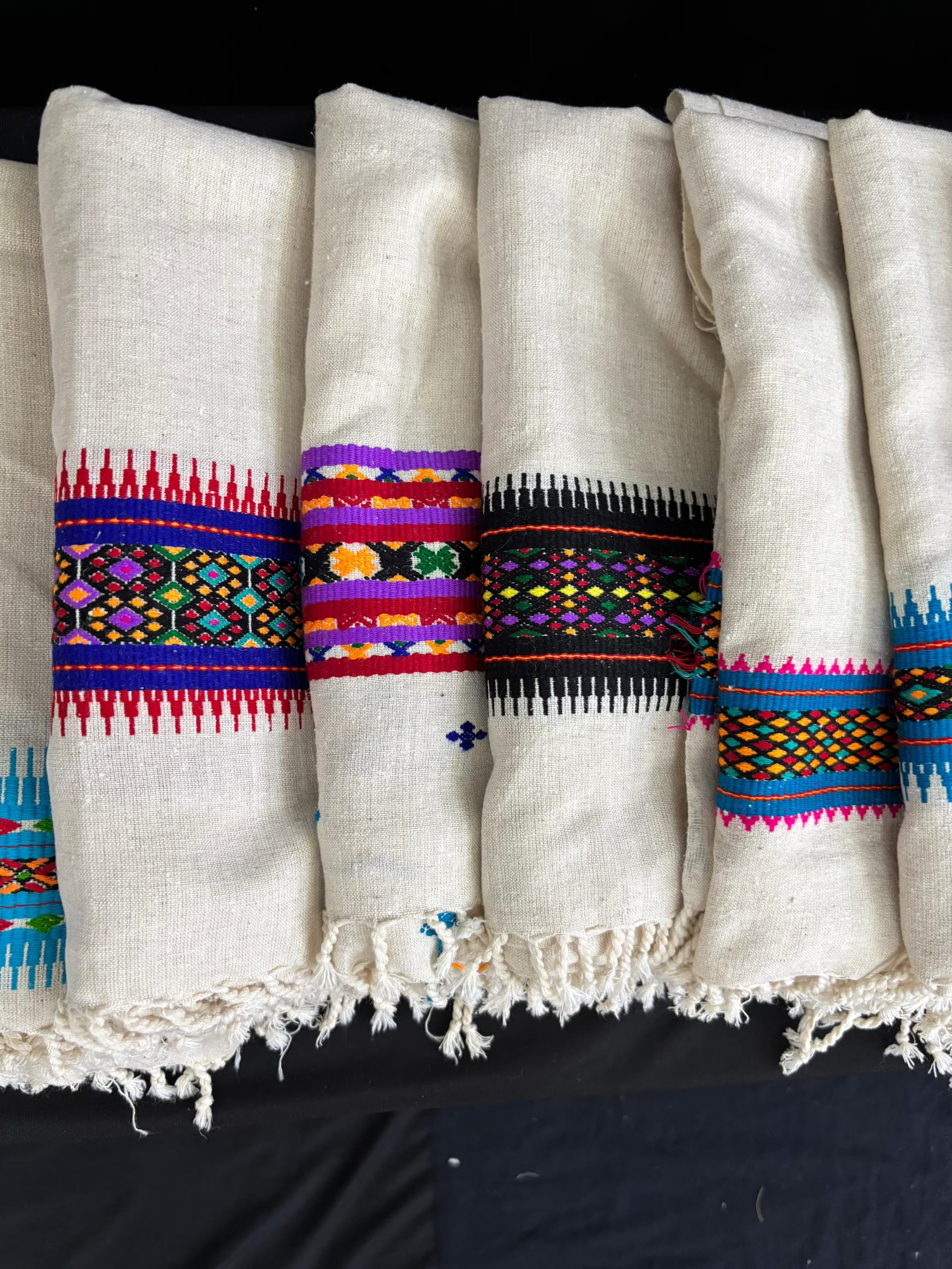 Folded cream eri-silk shawls laid in a row, each edged with a band of multicoloured geometric weaving in red, blue, purple, green and yellow, with knotted fringes