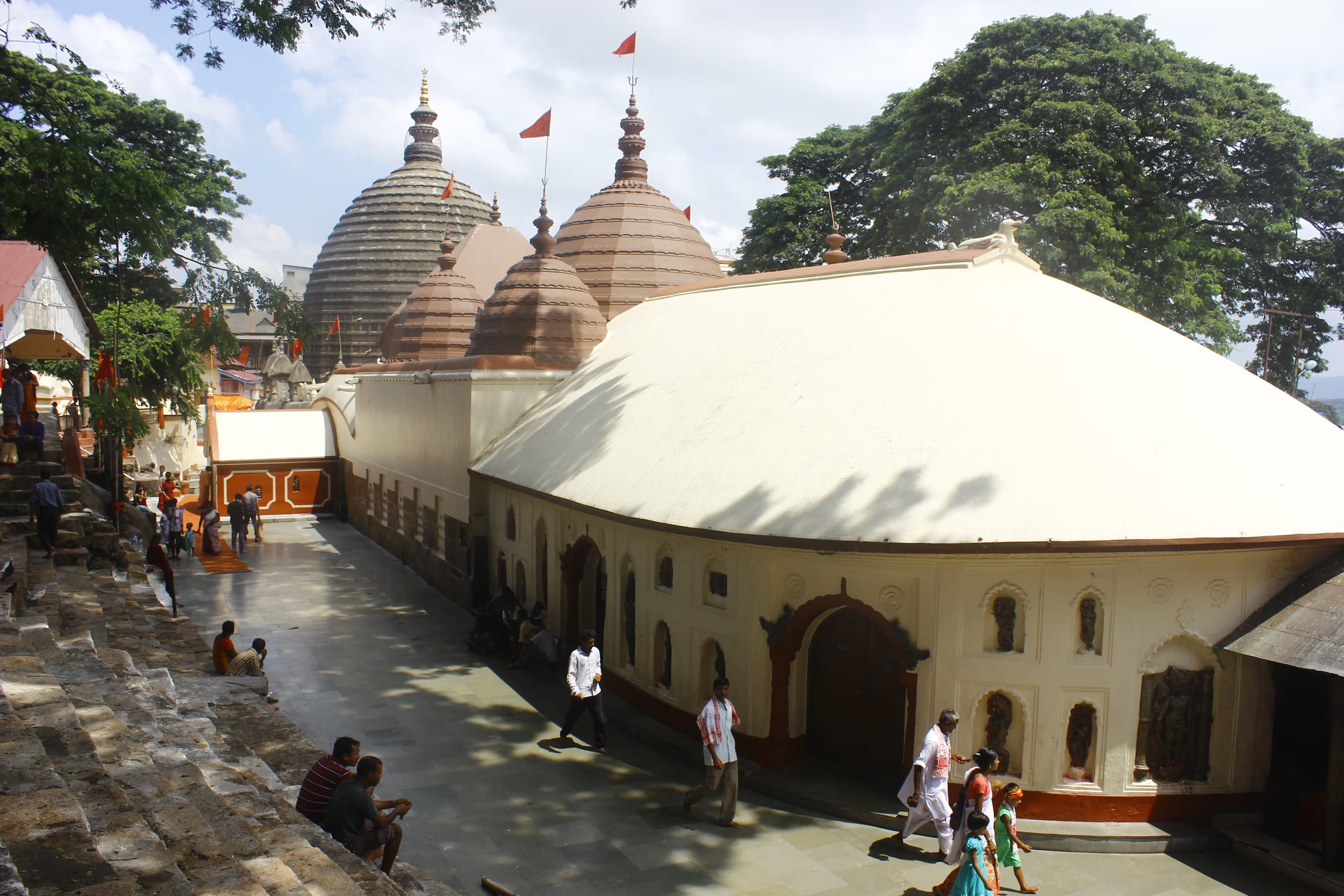 Elevated daytime view of the Kamakhya temple complex: a long whitewashed sanctum hall with a smooth rounded roof in the foreground and, behind it, a cluster of grey ribbed beehive-shaped shikharas topped with golden finials and saffron pennants; a stone-paved courtyard with a few small figures, green hills beyond under a bright sky