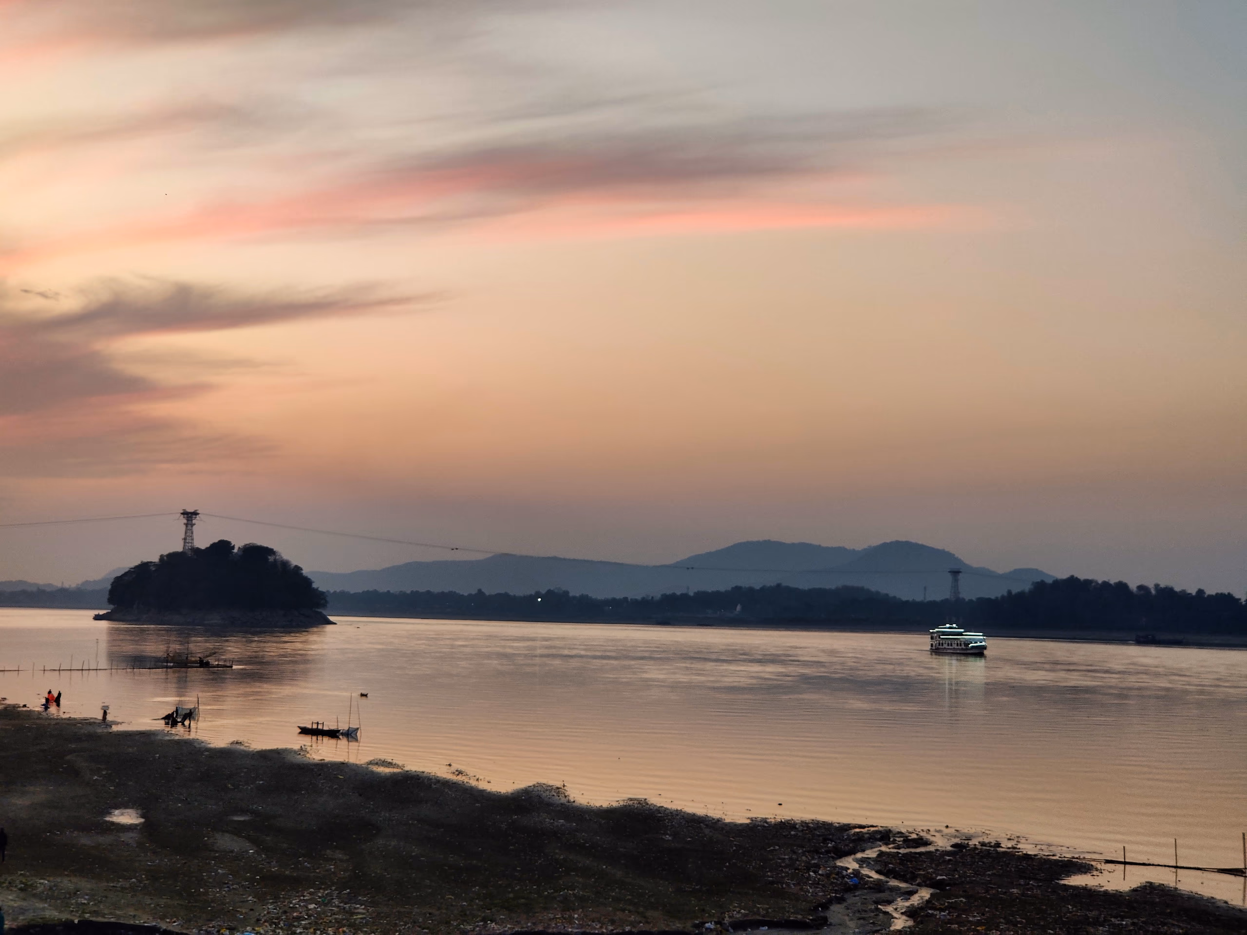 The broad Brahmaputra at sunset near Guwahati, with Umananda (Peacock Island) and its temple silhouetted at left and low hills along the far bank