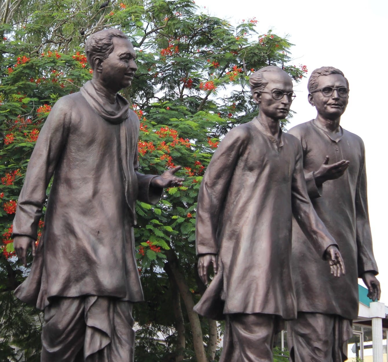 Three bronze full-figure statues of Assamese cultural figures, the central figure in round spectacles, beneath a flowering tree in Guwahati