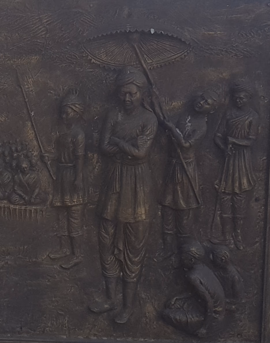 Bronze relief panel of Momai Tamuli Borbarua, the first Borbarua, standing beneath a ceremonial parasol with attendants