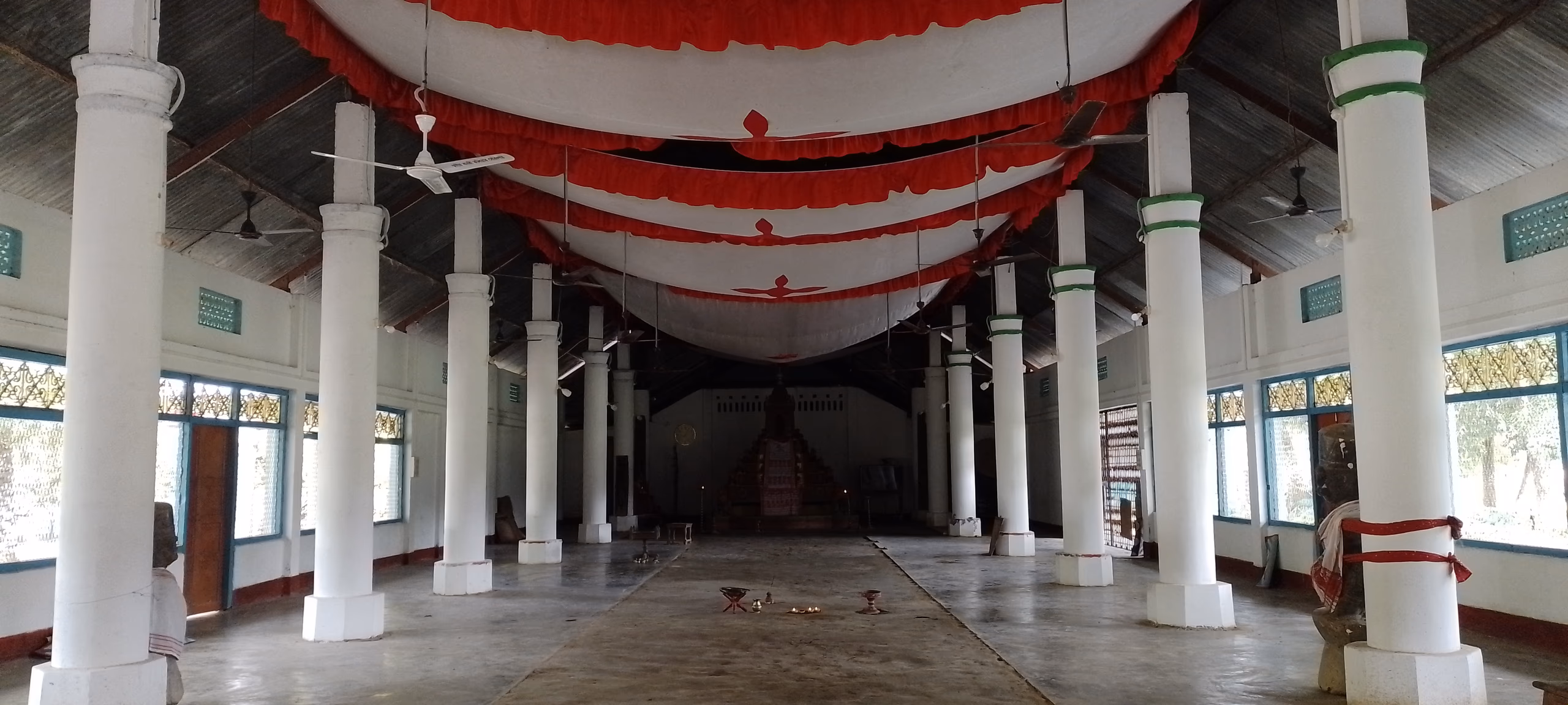 The long pillared prayer hall of the namghar at Bengenaati Sattra, with white columns, a red-and-white ceiling canopy, and the manikut shrine at the far end