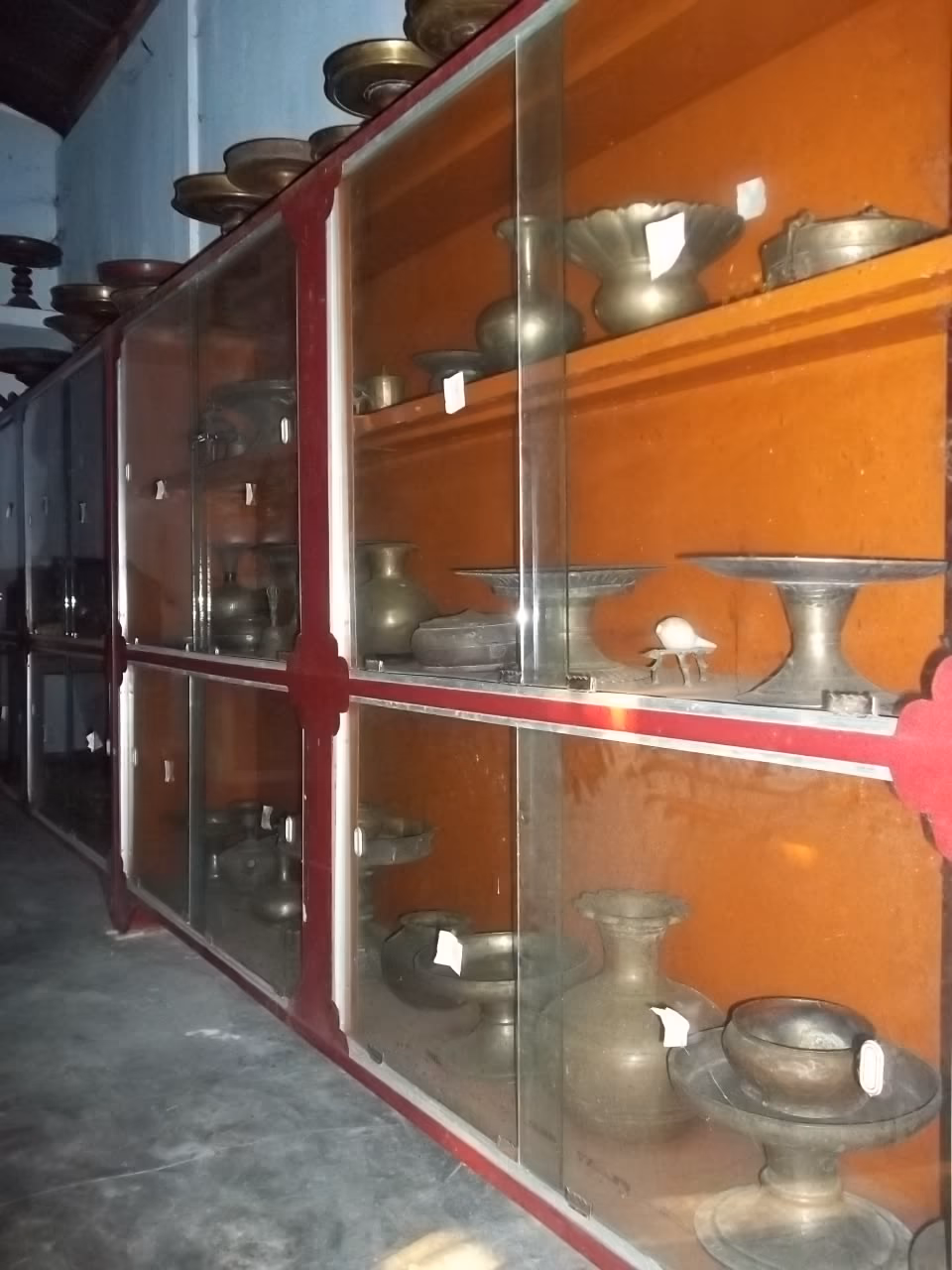 Glass display cases of brass and metal ritual vessels — bowls, footed trays, water pots — at Bengenaati Sattra