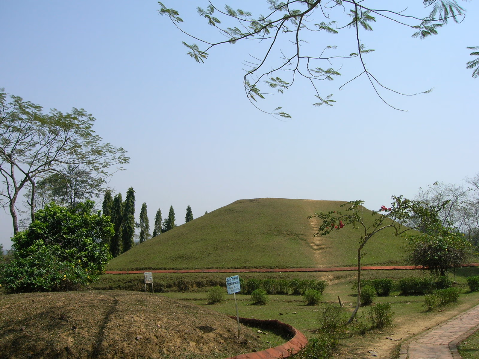 A large grass-covered hemispherical Ahom burial mound rising whole above the lawns at Charaideo, a narrow stepped path climbing its face, framed by trees and a low garden path