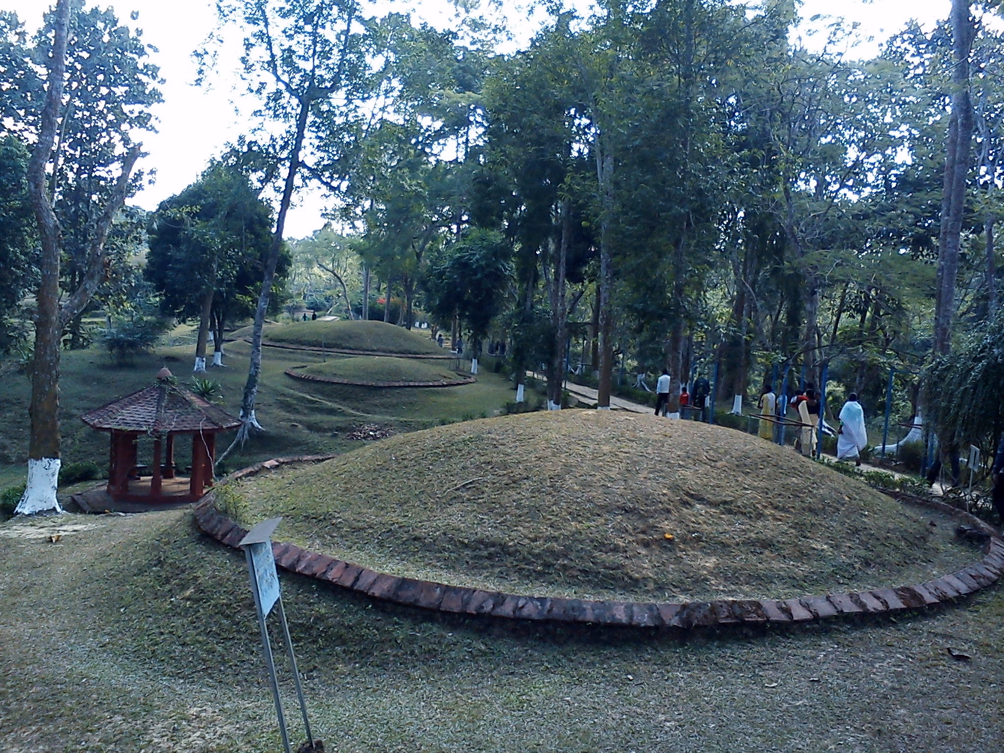 A grass-covered Ahom maidam at Charaideo ringed at its base by a low octagonal wall of red brick, a small white pavilion beside it and further green mounds rising along the wooded ridge behind