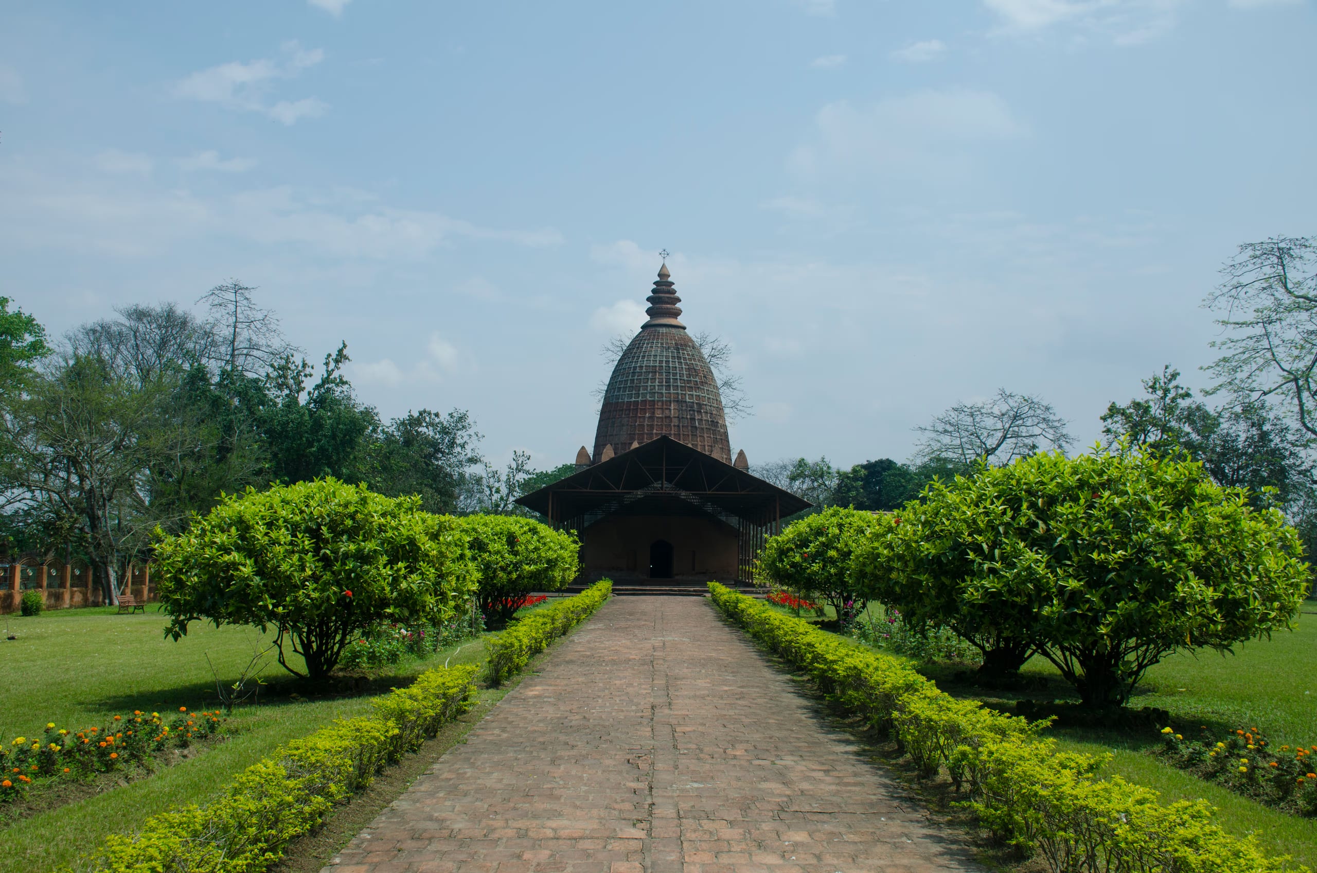 The brick Joydol at Joysagar seen straight down a paved walkway flanked by clipped hedges and flowering shrubs on green lawns, the tall ribbed dol tower rising behind its pillared hall under a bright sky