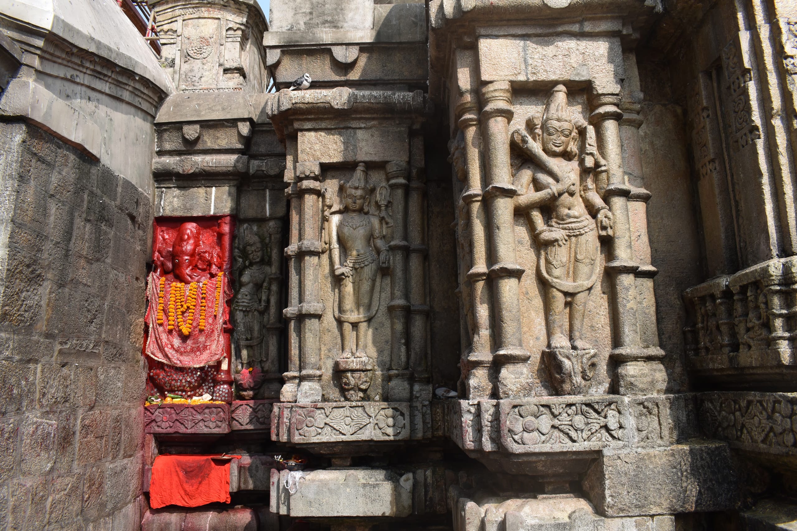 Weathered grey stone panels set into the temple wall: a slender standing deity in a niche framed by colonnettes, a fuller multi-armed figure beside it, and a vermilion-smeared image draped in marigold garlands at left