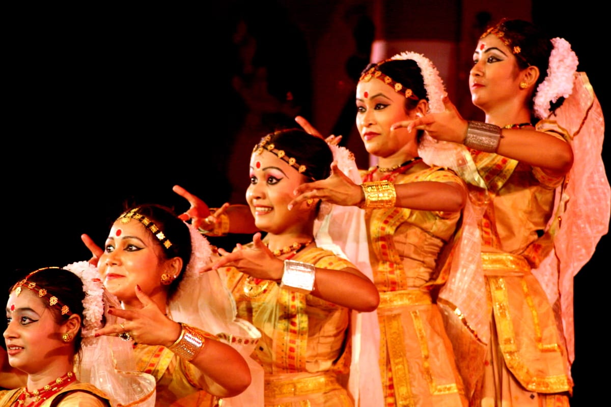 A row of Sattriya dancers in white-and-gold costume holding a synchronised hand gesture, with the white chador, jewellery and red-dotted brow of the form