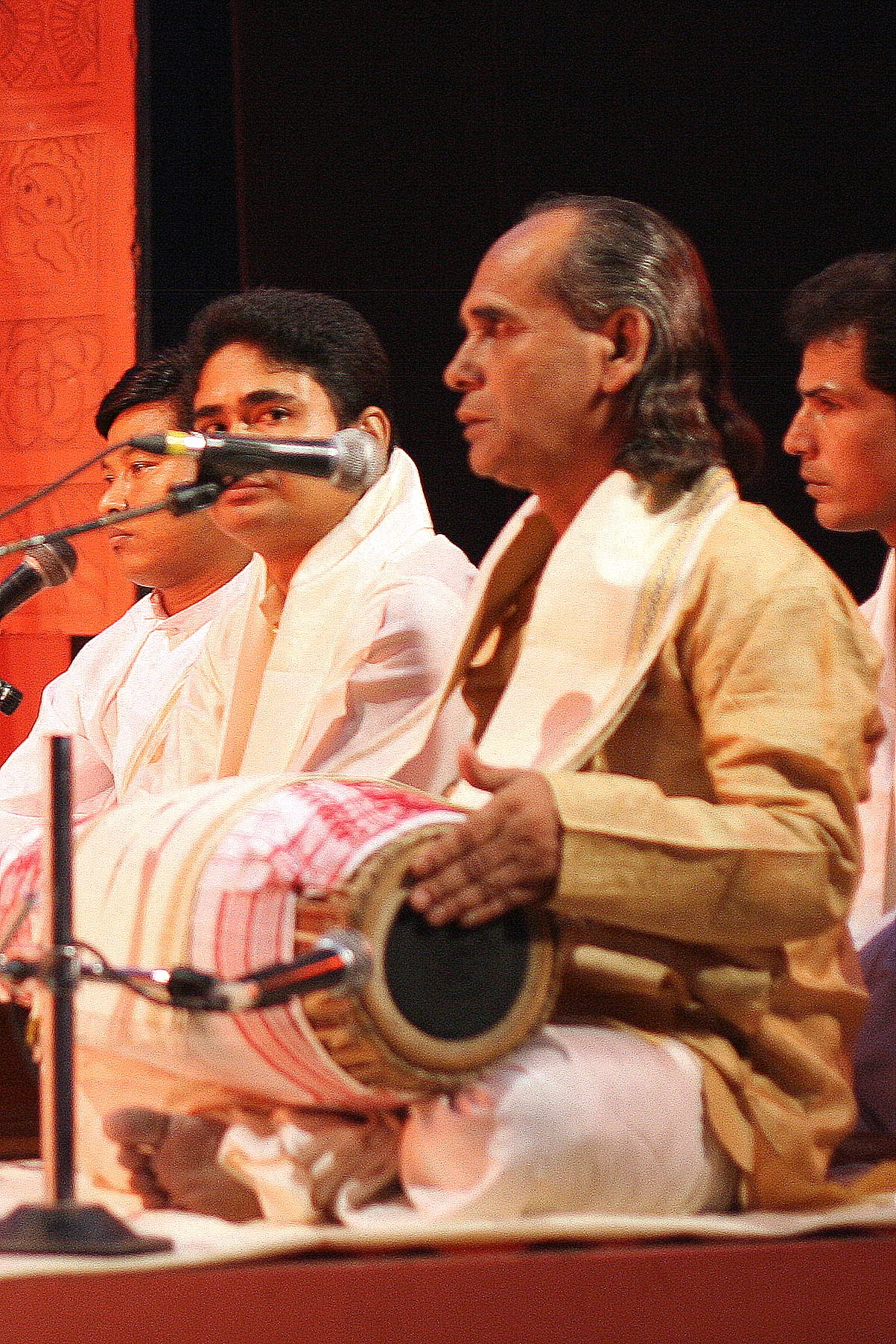 An older man in cream dress with a khol drum across his lap, singing among seated musicians at microphones on a stage