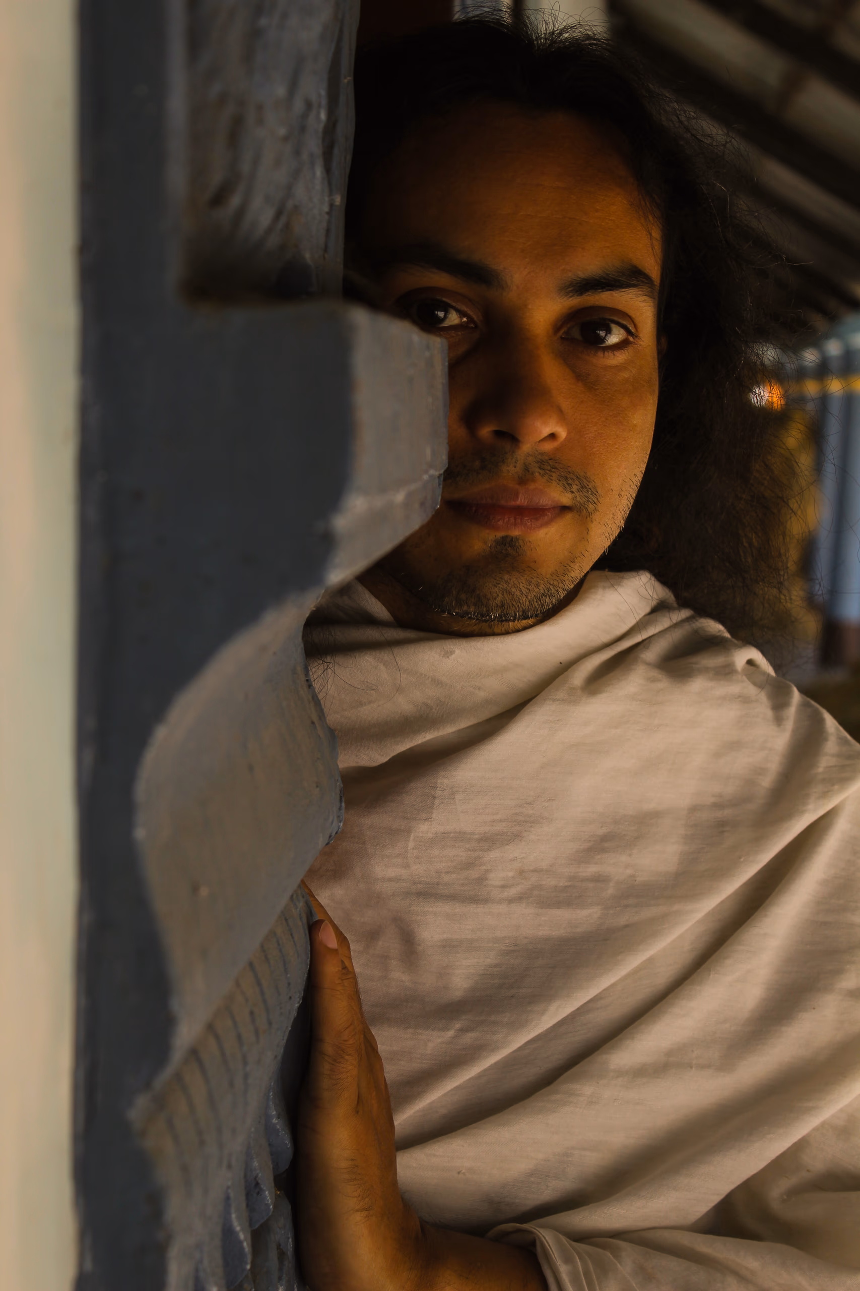 A young monk wrapped in a plain white cloth leans against a painted blue doorframe of the monastery, lit by warm low-angle light, looking calmly toward the camera