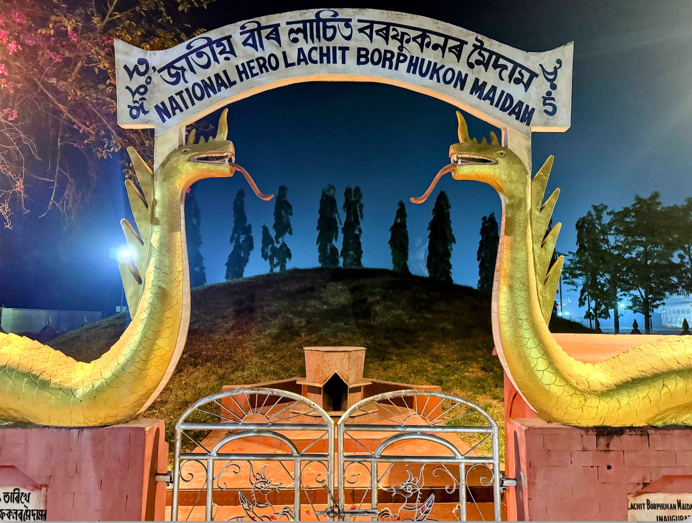The ornamental gateway to the Lachit Borphukan Maidam at night: twin golden serpent-dragon arms curve up to a painted signboard reading National Hero Lachit Borphukan Maidan in Assamese and English, an iron gate below, the grassed burial mound and tall trees floodlit behind