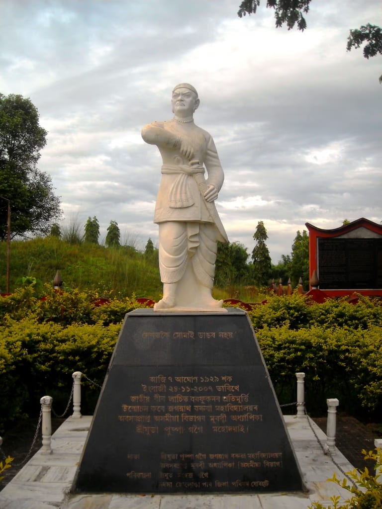 A tall white statue of Lachit Borphukan on a black inscribed plinth, the Ahom commander standing in a long robe with one arm raised, set in a landscaped garden with the green burial mound and trees behind under a cloudy sky