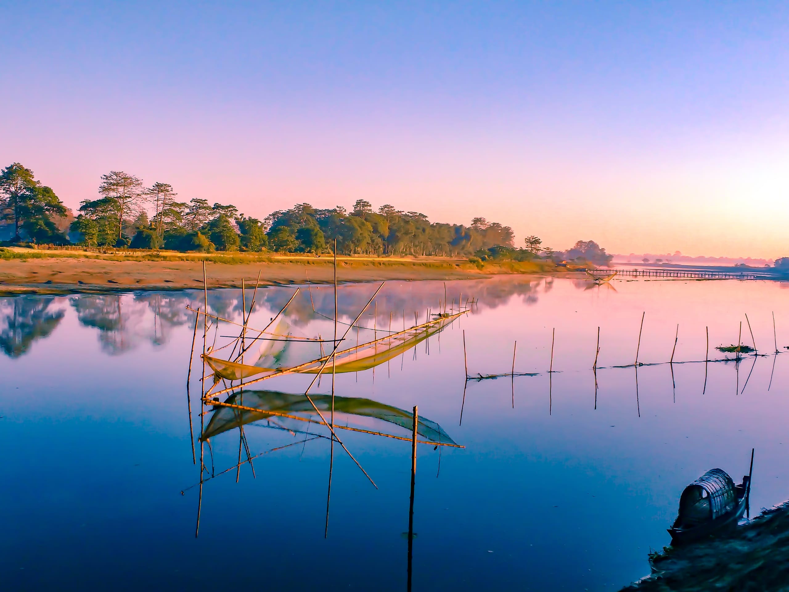 A still wetland channel on Majuli at first light, bamboo fishing-net frames standing in the glassy water before a low tree-lined island bank, a country boat at right