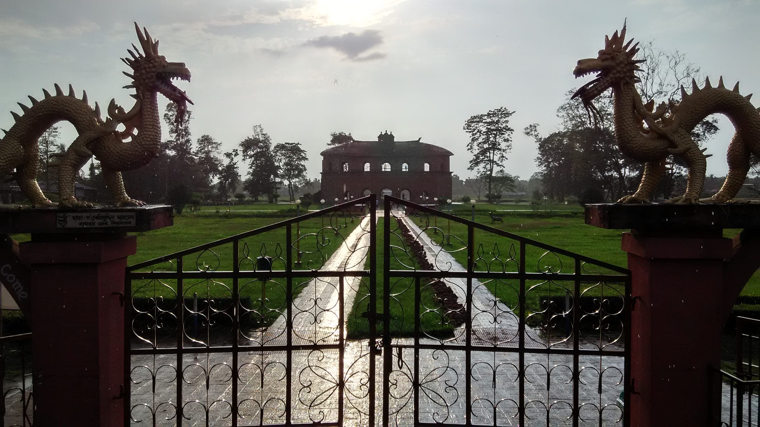 The Rang Ghar seen down a long axial garden, framed by a pair of golden dragon statues mounted on the gateposts above a wrought-iron gate, the oval pavilion centered in the distance