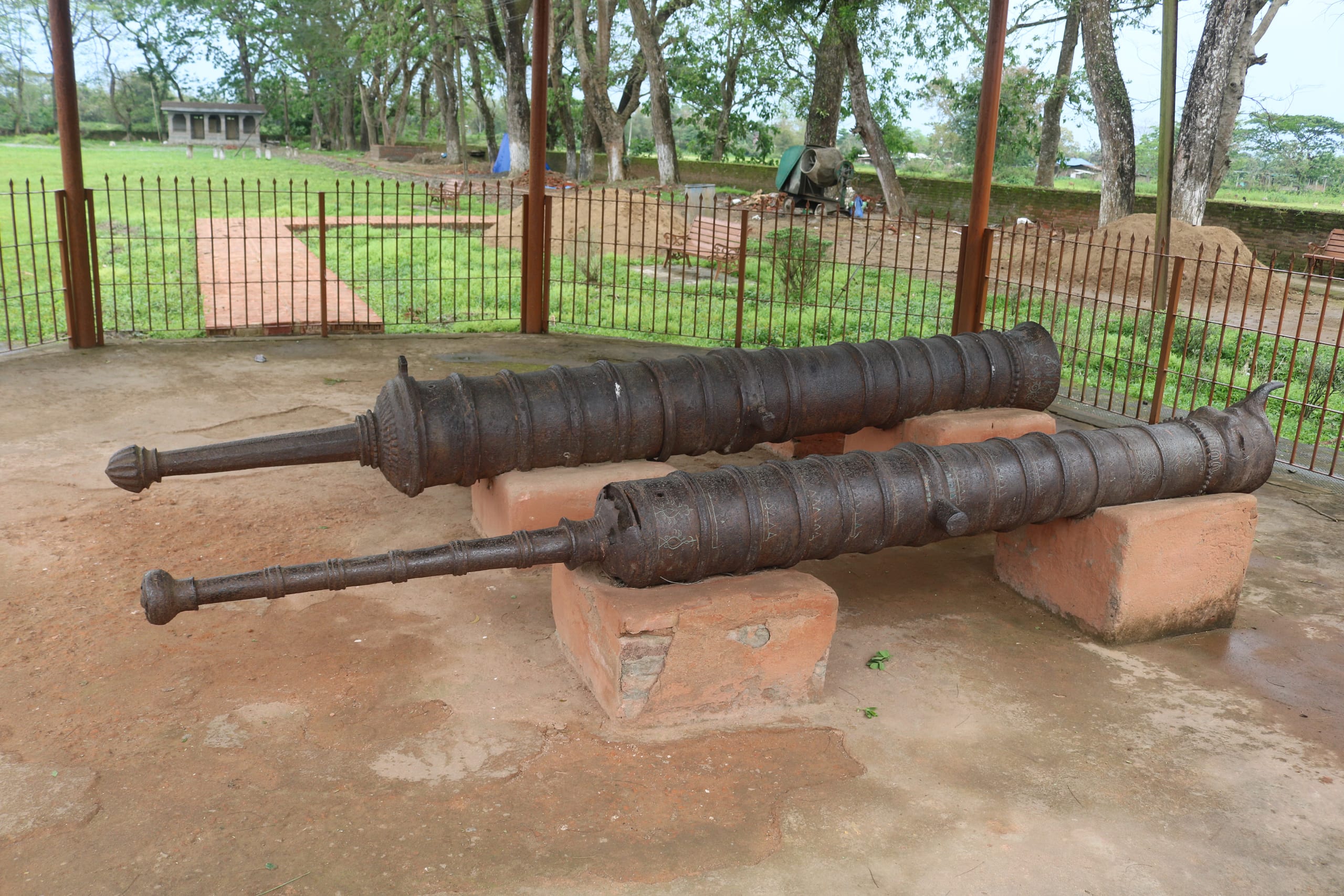 Two long iron Ahom-period cannons resting on brick plinths under an open shelter on the Talatal Ghar grounds