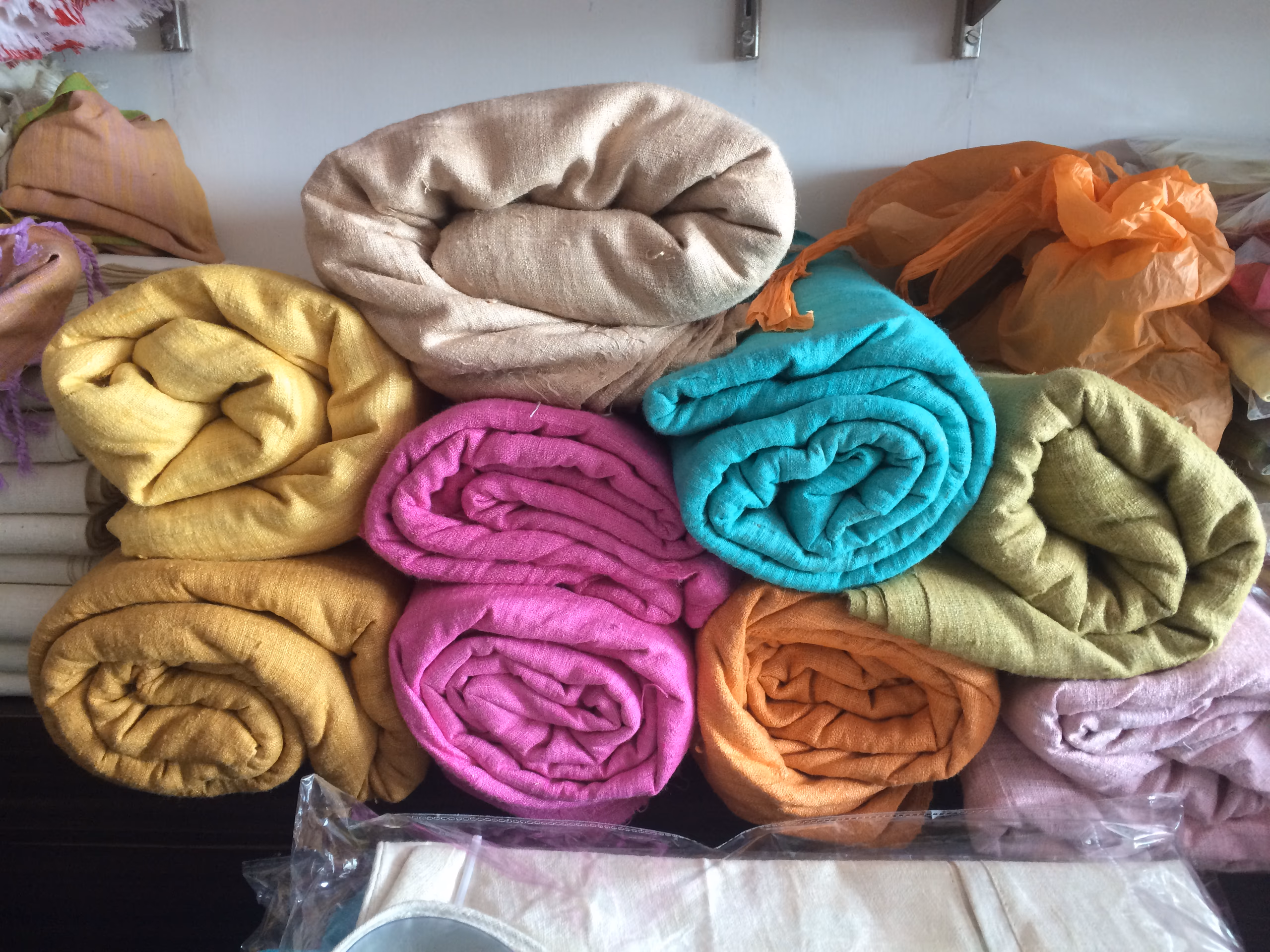 Rolled bolts of eri silk fabric in cream, mustard, pink, teal, ochre and green stacked on a shelf