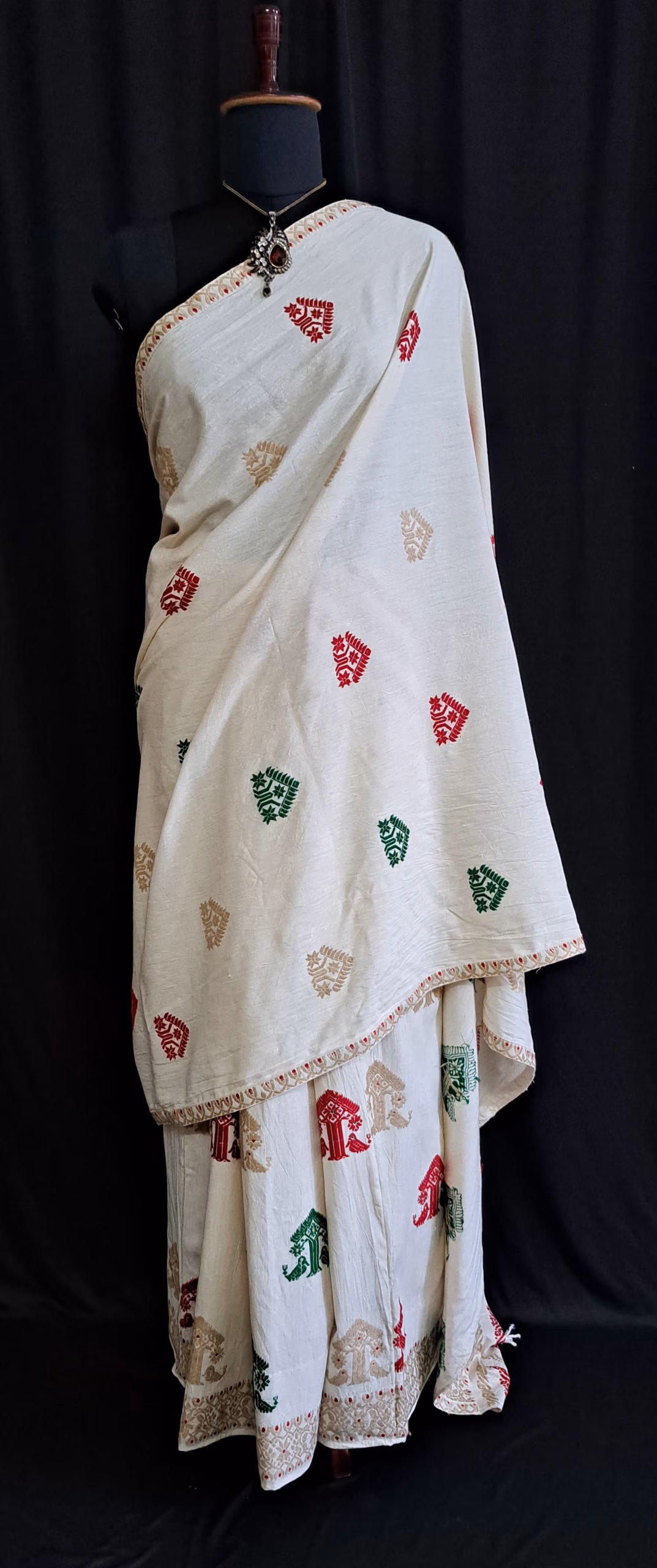 An ivory-white Assamese mekhela sador draped on a stand, its white field scattered with small woven motifs in red, green and gold