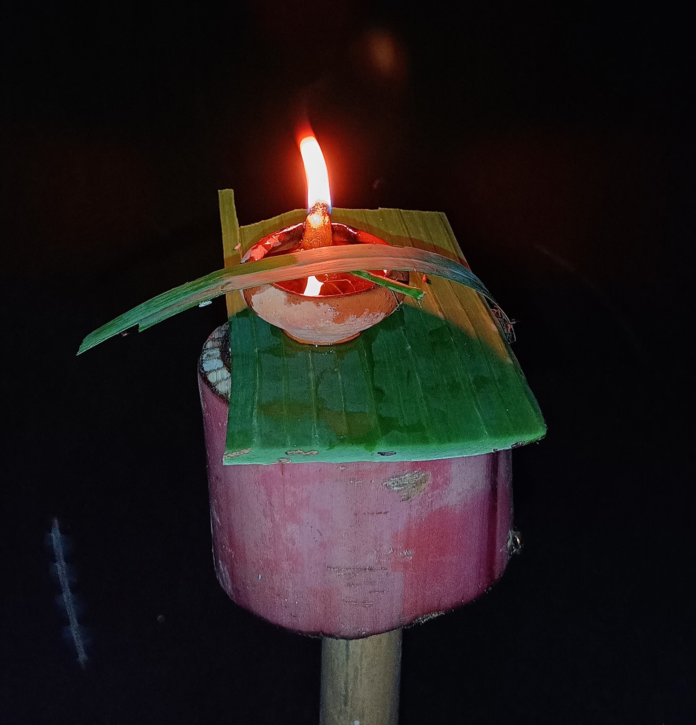An earthen lamp burning atop a bamboo pole, sheltered by a folded banana-leaf hood, against the night