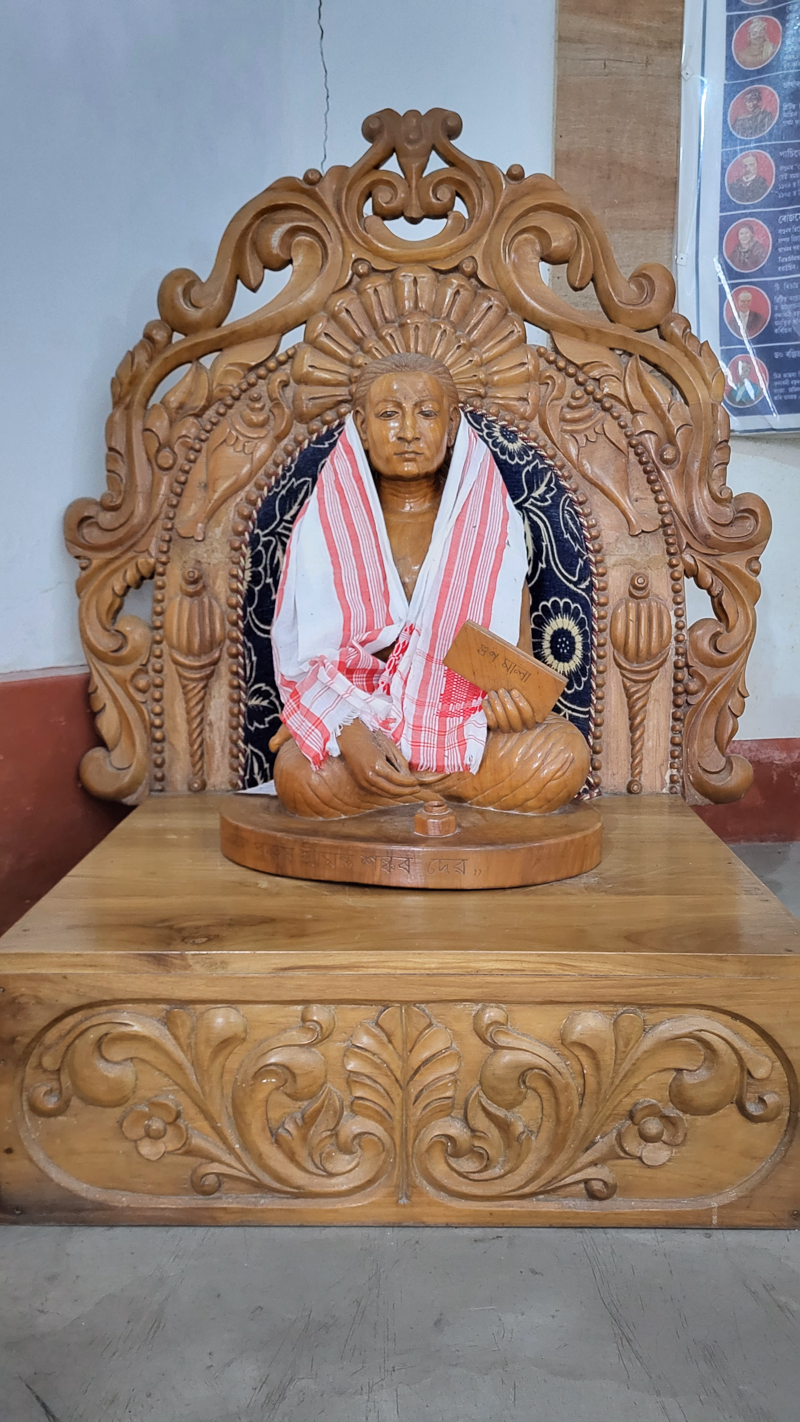 Carved wooden statue of Srimanta Sankardev seated cross-legged on a lotus pedestal within an ornate wooden halo-frame, draped in a red-and-white Assamese gamosa and holding a small inscribed manuscript