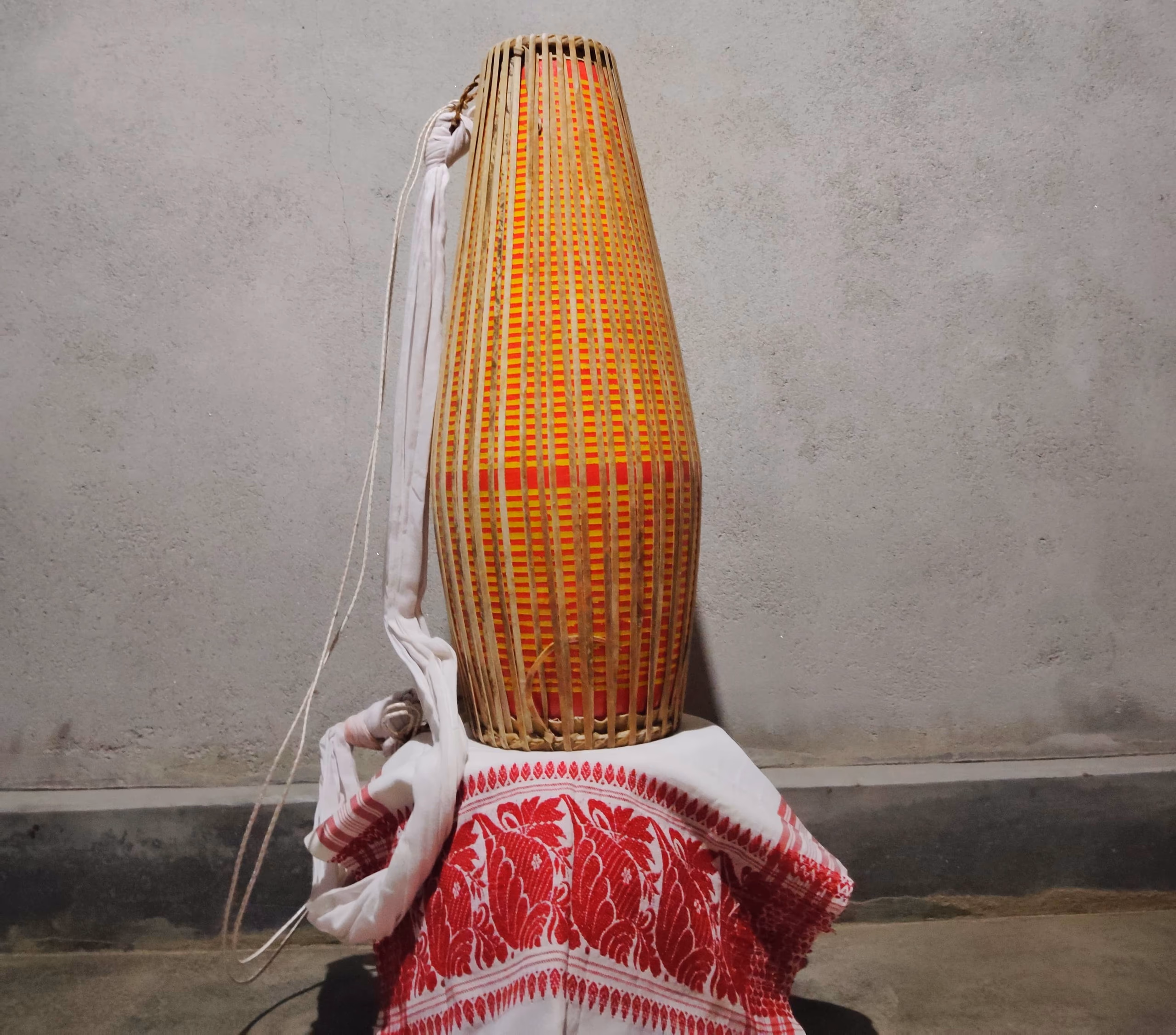A single two-headed khol drum standing upright on its narrower head, its long tapering body wrapped in close vertical bamboo-and-cane lacing over orange and red banding, a white cloth carrying strap looped down one side, the drum resting on a folded red-and-white gamosa against a plain grey wall
