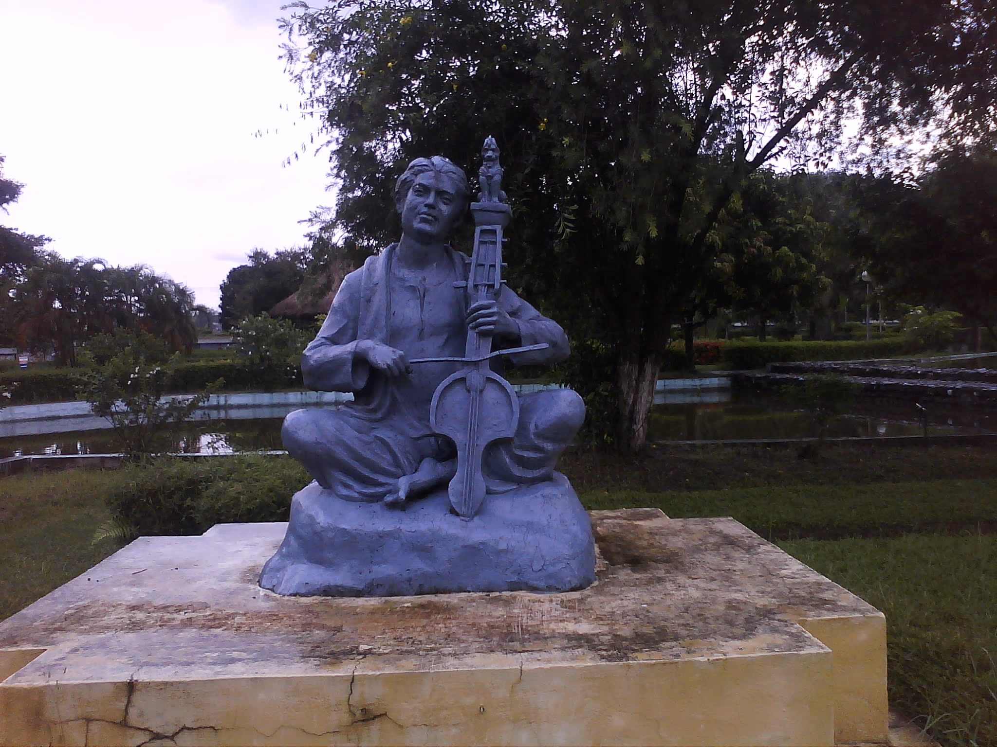 A grey stone seated statue of Sankardev cross-legged on a plinth, playing a long-necked stringed instrument held upright, with trees and a pond behind