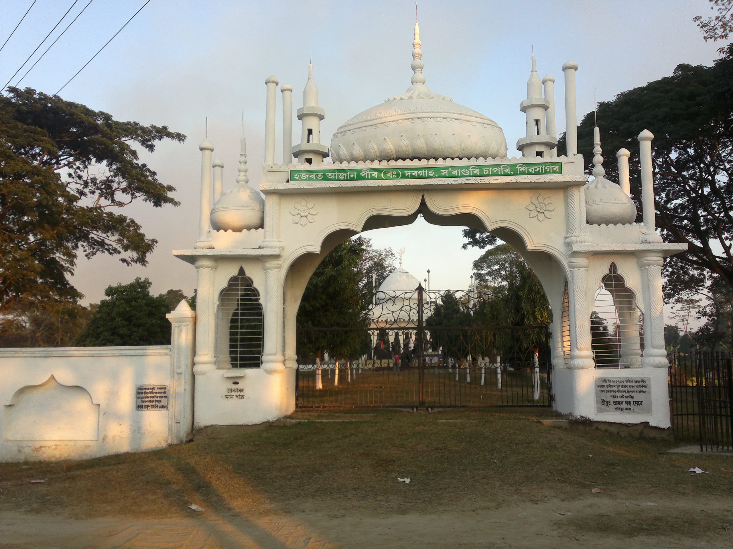 Whitewashed Indo-Islamic gateway arch with small domes and minaret-like finials, an Assamese-script name board across the arch, the domed shrine visible through it down a path