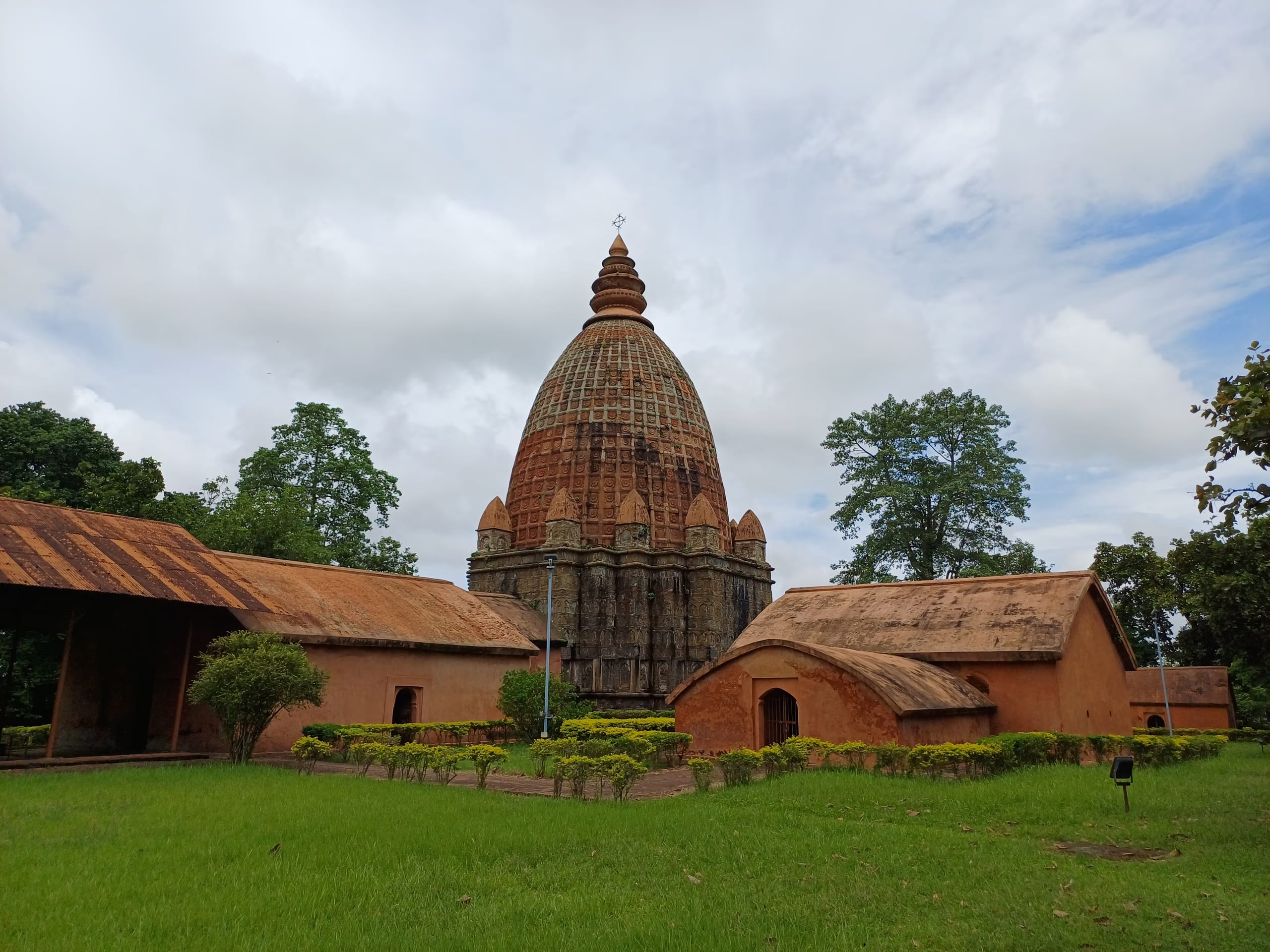 A tall brick temple with a ribbed, curvilinear spire rising over lower red-brick outbuildings with curved roofs, set behind a mown lawn and hedge under a cloudy sky
