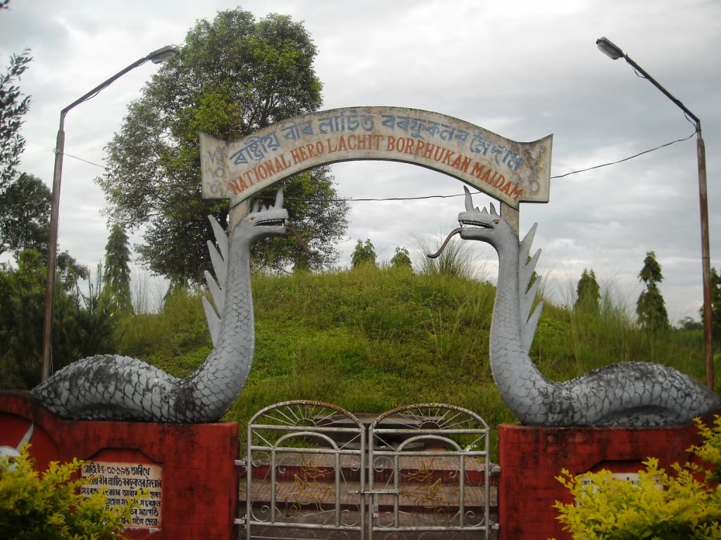 Grass-covered burial mound behind an arched gateway flanked by two serpent sculptures, signed National Hero Lachit Borphukan Maidam