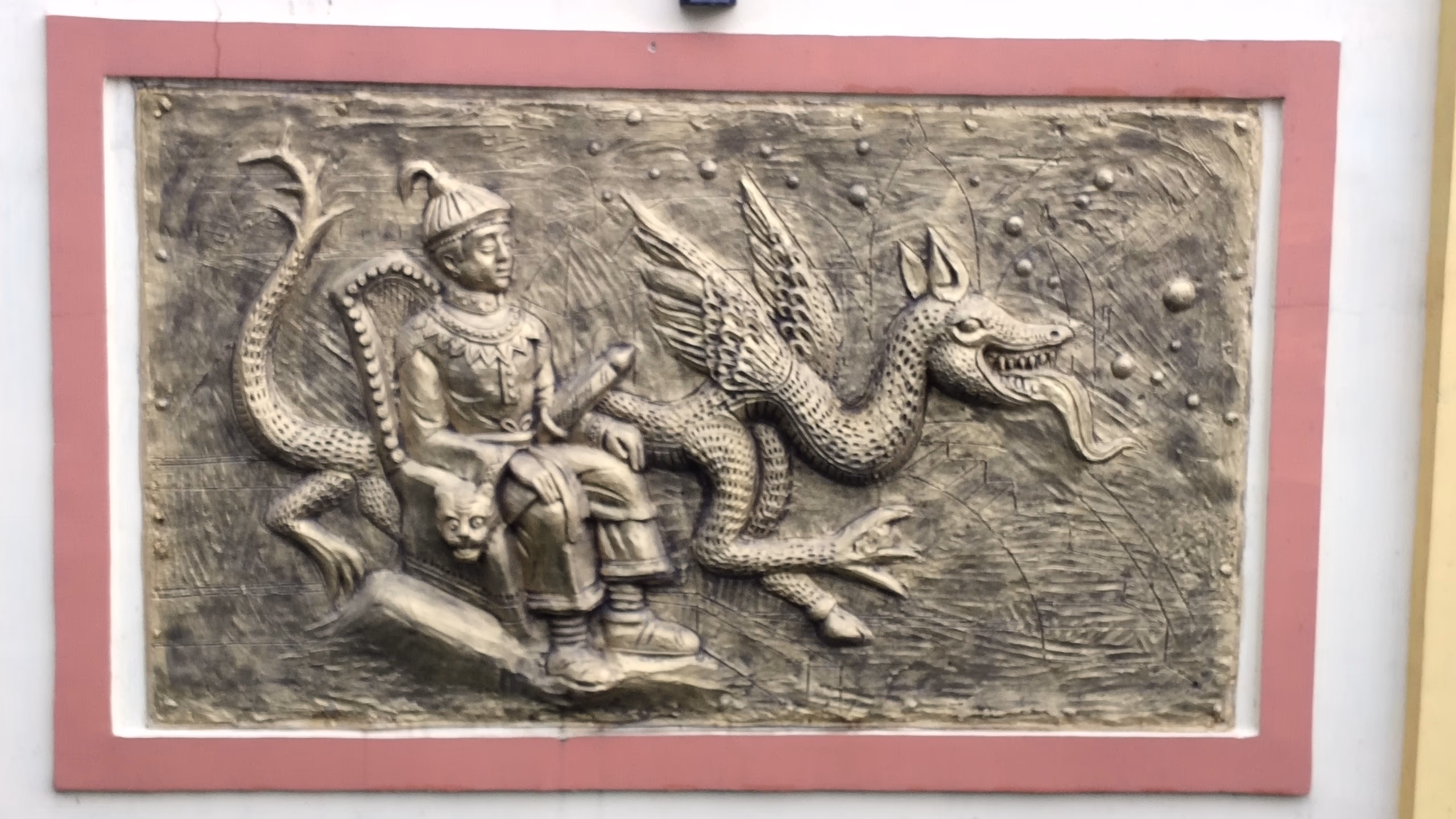 Cast relief panel showing Sukaphaa enthroned in a conical Tai cap, beside a winged dragon, set in a pink-bordered frame on a wall