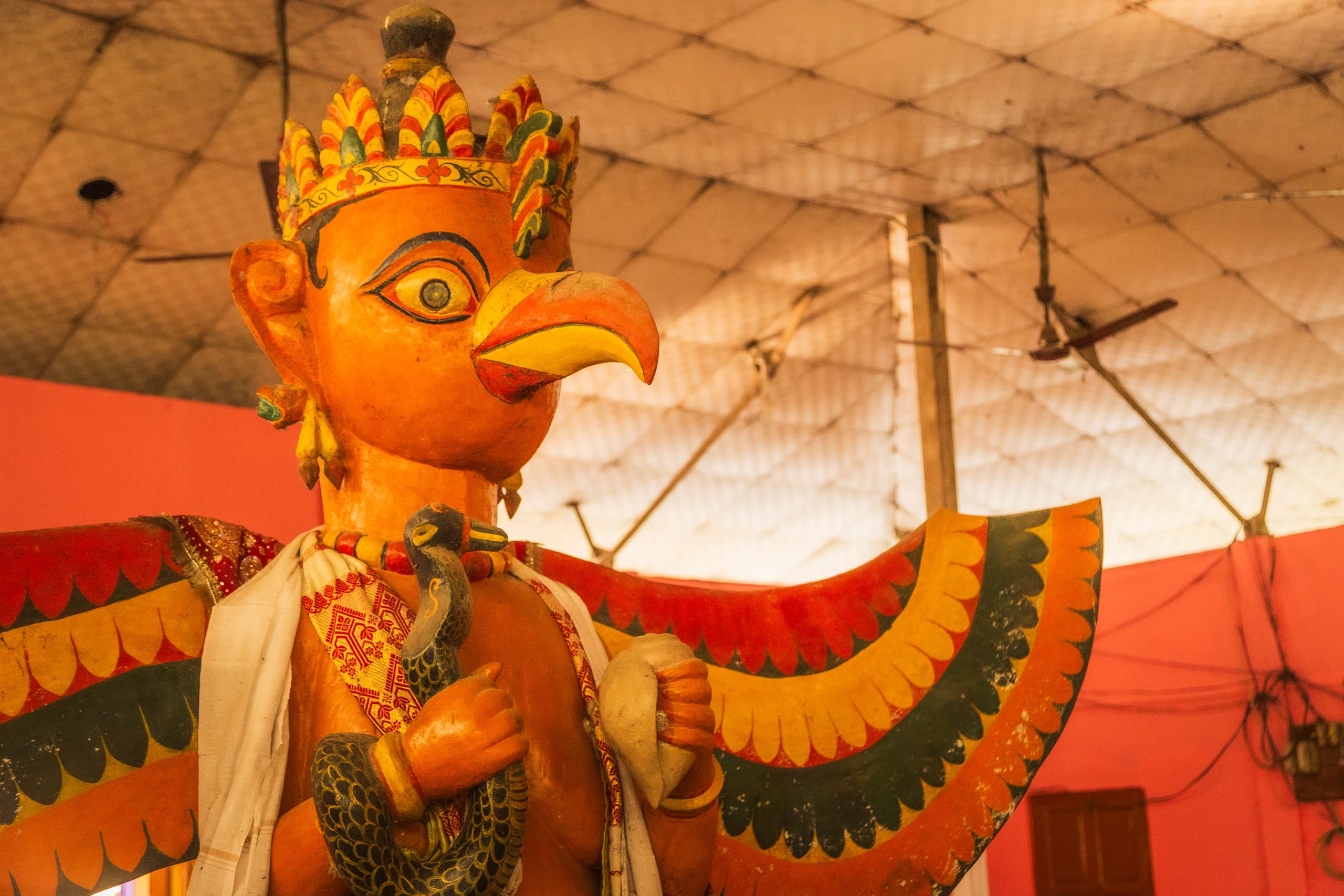 A large brightly painted effigy of Garuda with a crowned eagle head and outstretched green-and-red wings, raised under a festival canopy at Auniati Sattra