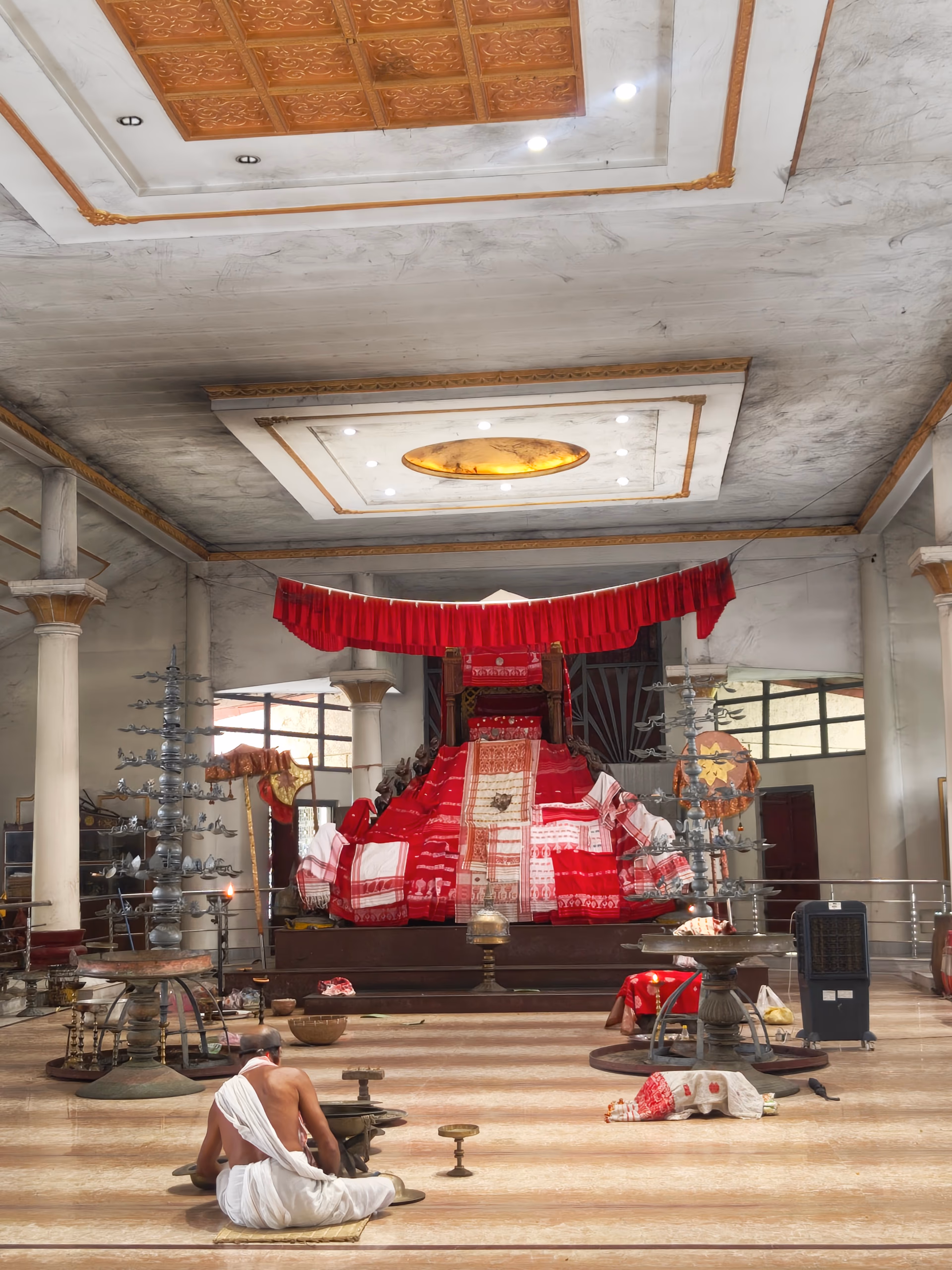 The marble-floored prayer hall of the Batadrava Than, with a red-draped throne bearing the sacred manuscript at its centre and brass ceremonial lamps to either side