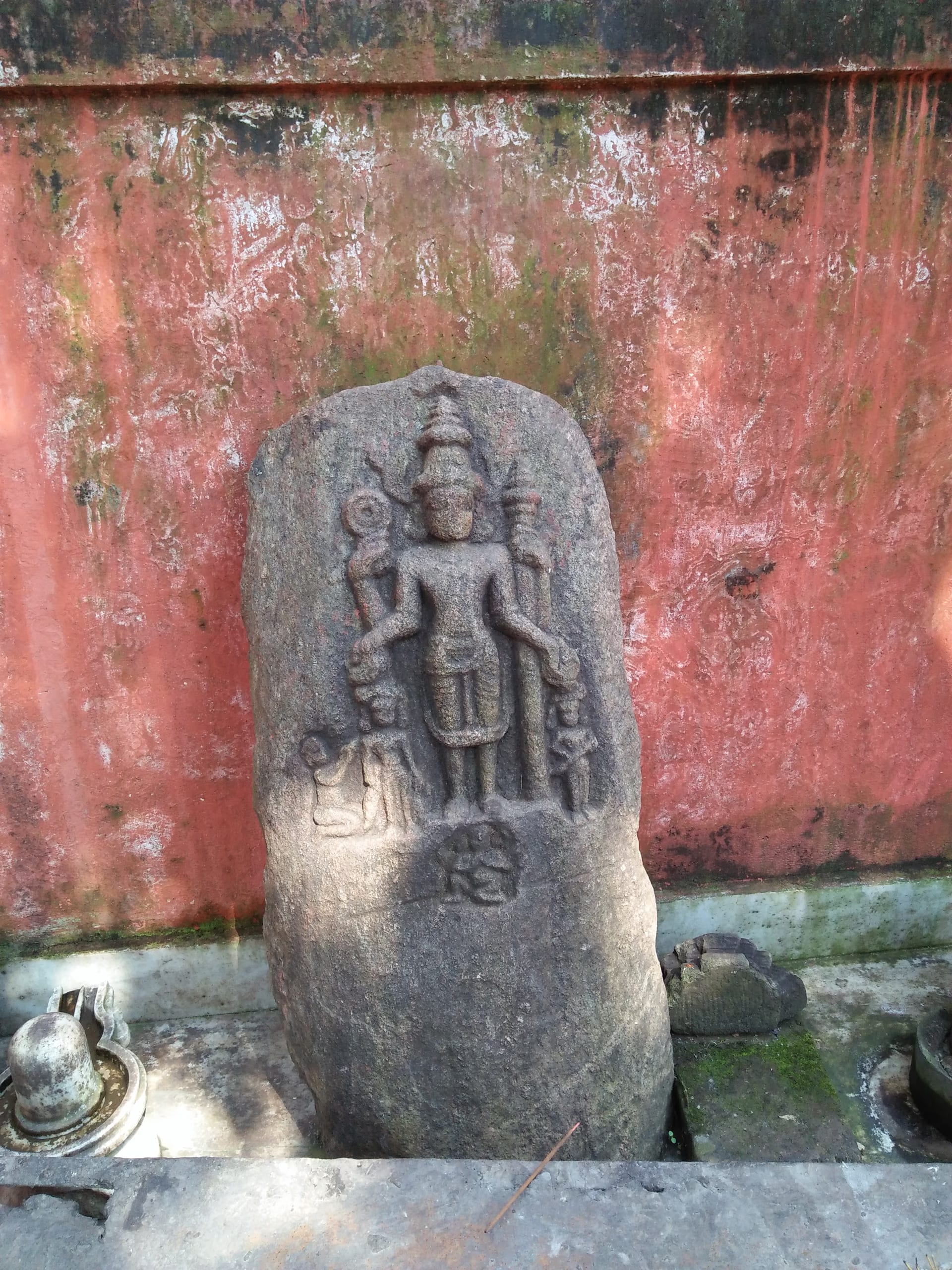 A weathered grey stone stele of a four-armed standing deity, set against a red temple wall at Basistha, Guwahati, with a small bell and stone fragments at its base