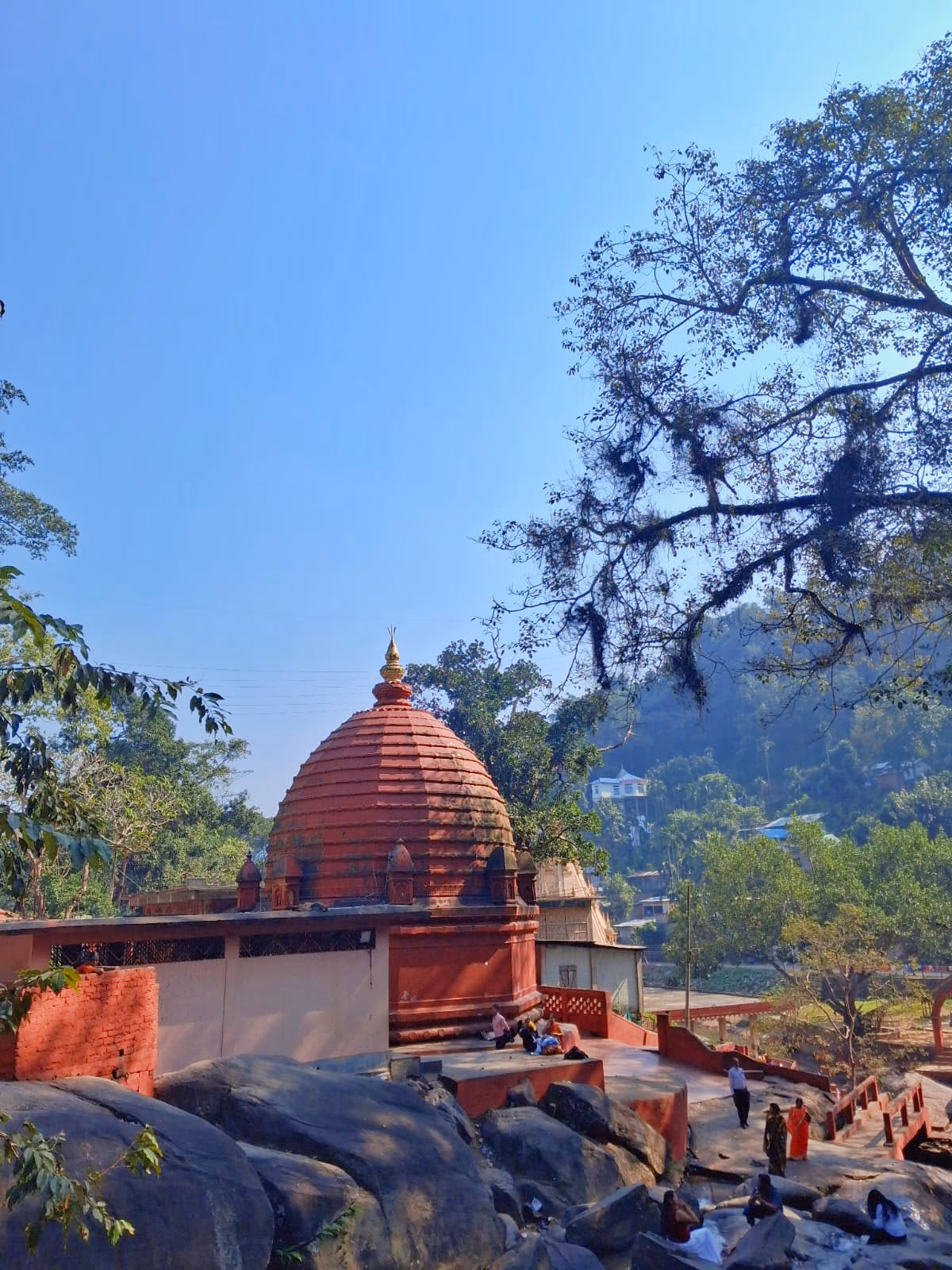 The Basistha temple complex with its ribbed orange-red sikhara set among large boulders, with forested hills rising behind under a clear blue sky