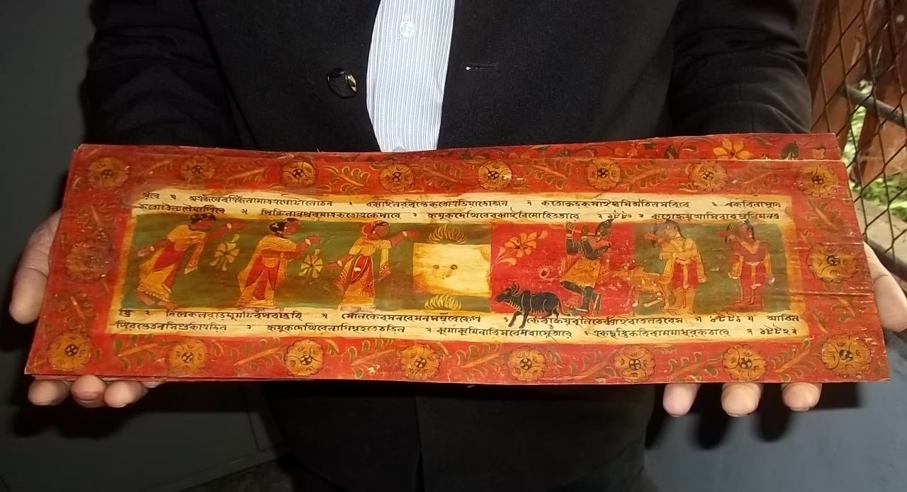A long illustrated manuscript leaf with rows of Assamese script and painted miniatures of figures and animals within red floral borders, held open for view