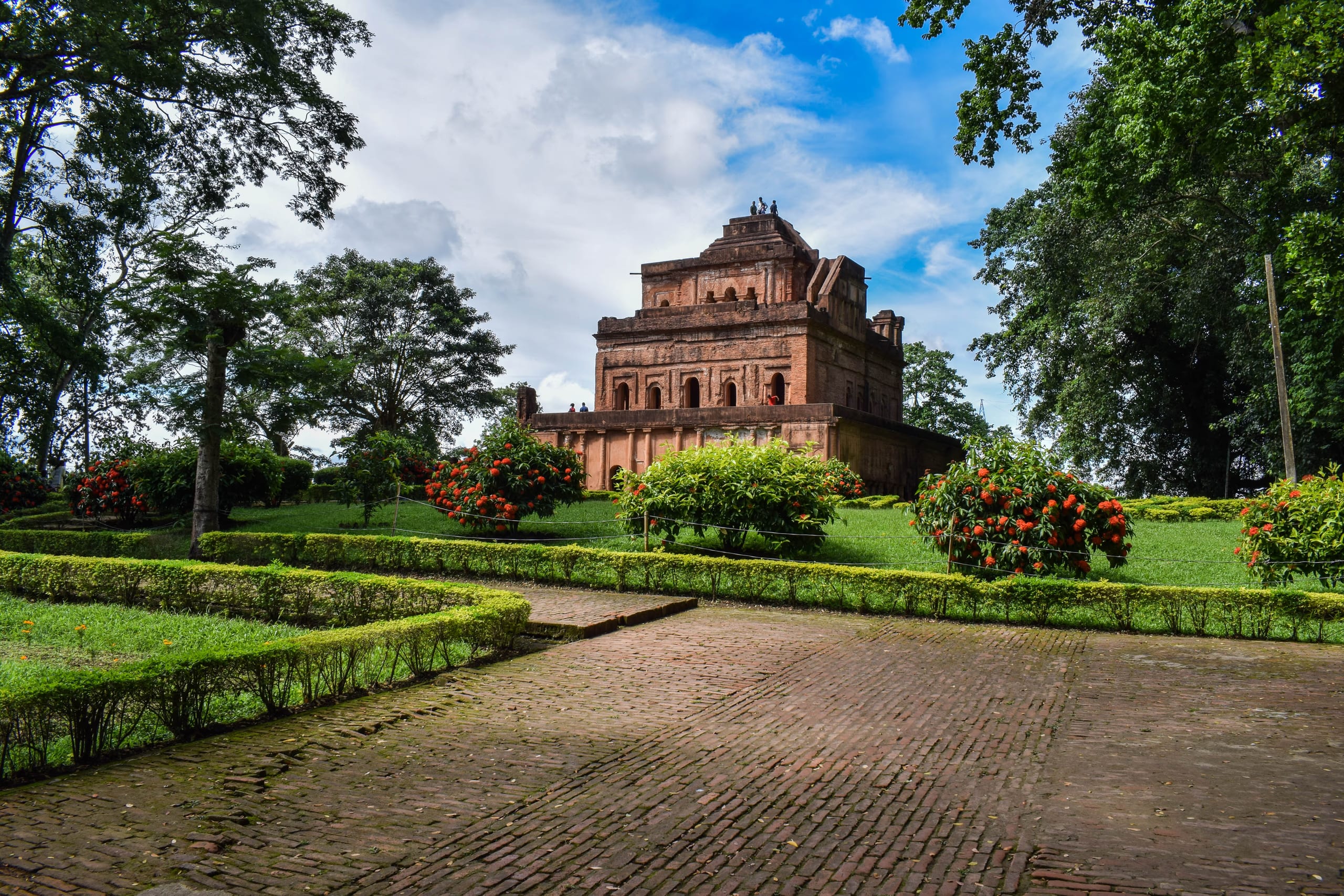 Three-quarter view of the Kareng Ghar at Garhgaon, the tiered brick Ahom palace seen from the front-left across lawns and flowering shrubs, brick paths converging toward it under a cloud-streaked sky