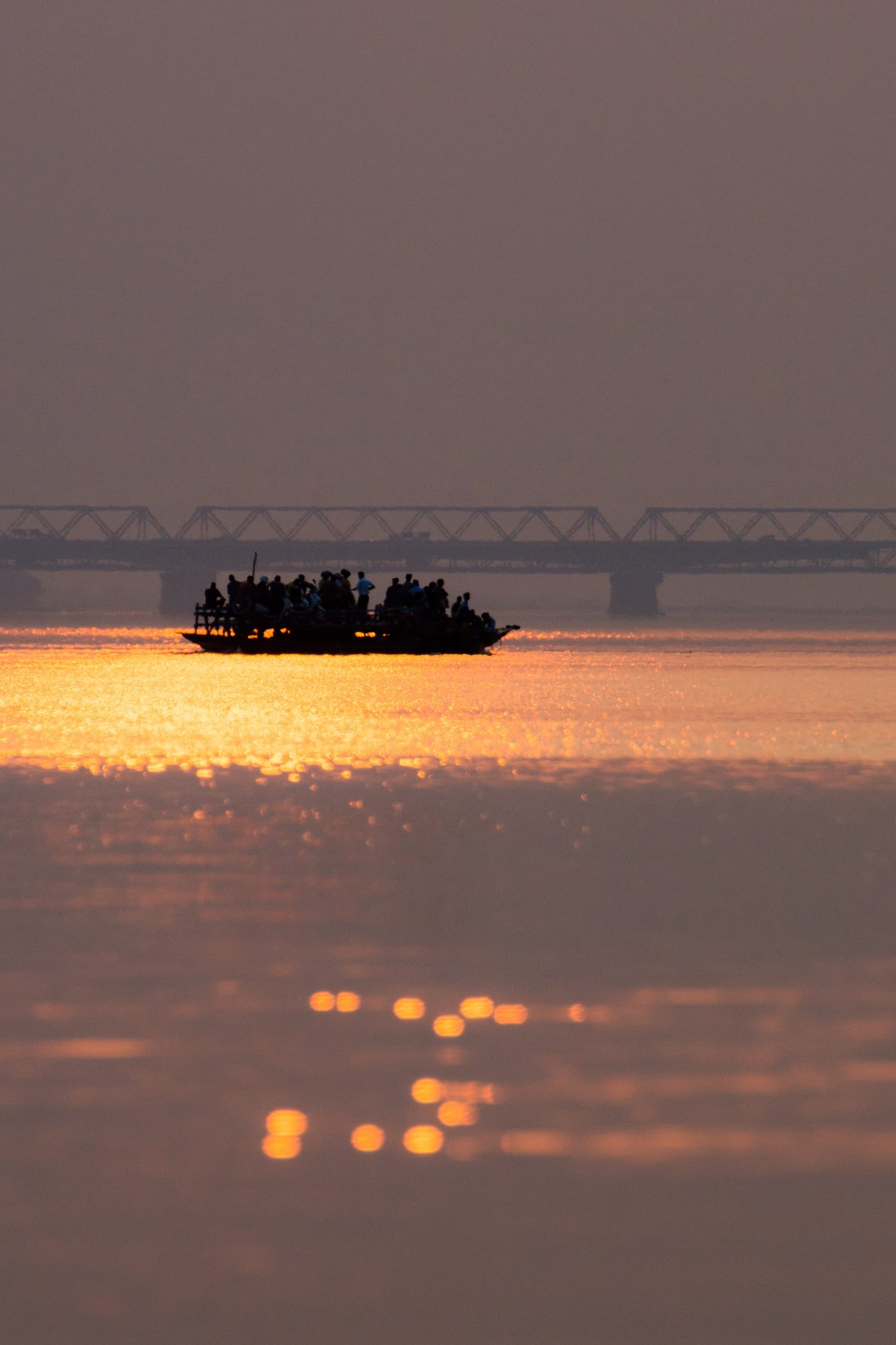 A crowded country ferry boat in silhouette crossing the Brahmaputra at sunset, the broad river a sheet of molten gold, the Saraighat bridge a faint silhouette on the misty horizon behind