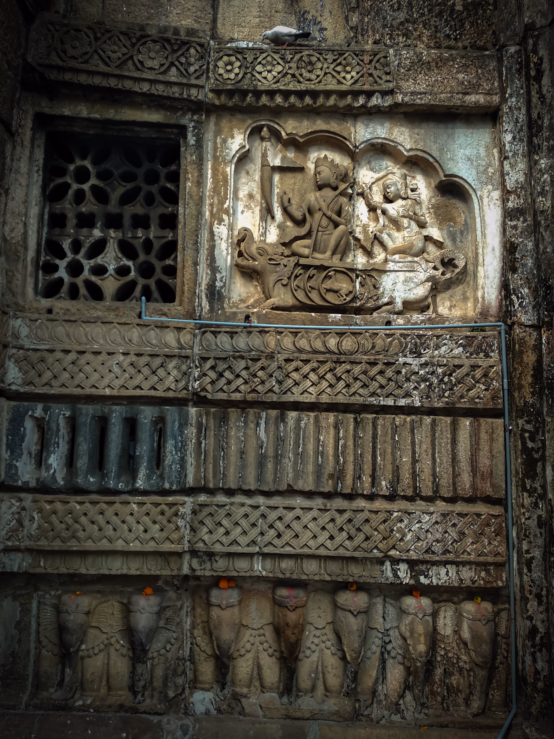 Carved stone panels on the wall of the Hayagriva Madhava temple at Hajo: a perforated lattice window, a figural relief in an arched niche, geometric friezes, and a row of carved elephants along the base