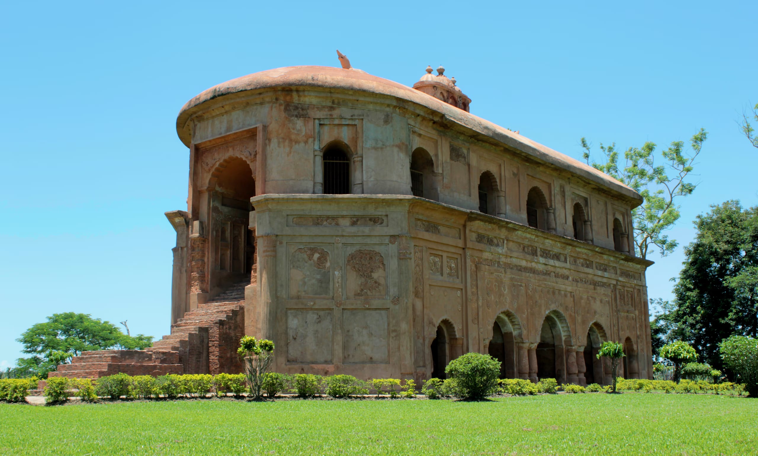 Three-quarter view of the Rang Ghar against a clear blue sky, showing its curved oval end, carved side wall and boat-shaped roof above a green lawn