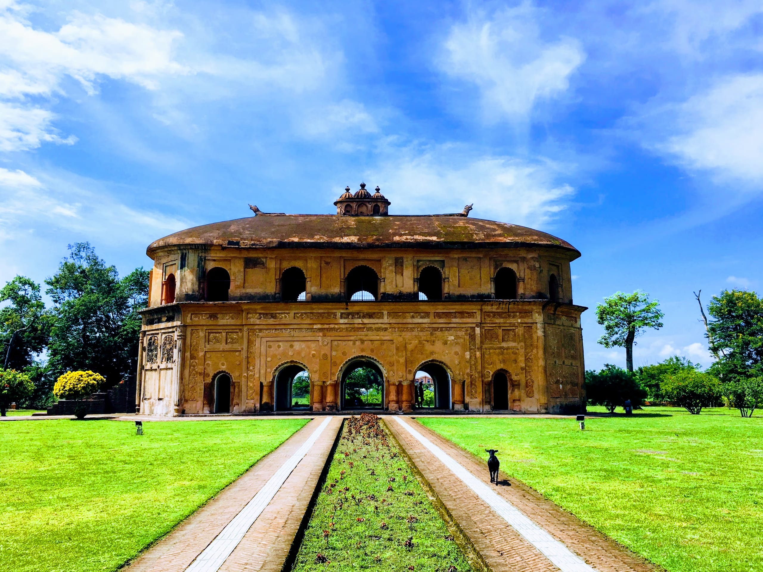 Frontal view of the Rang Ghar under a dramatic blue sky with sculptural clouds, the central garden path leading to the arched lower storey across a bright green lawn