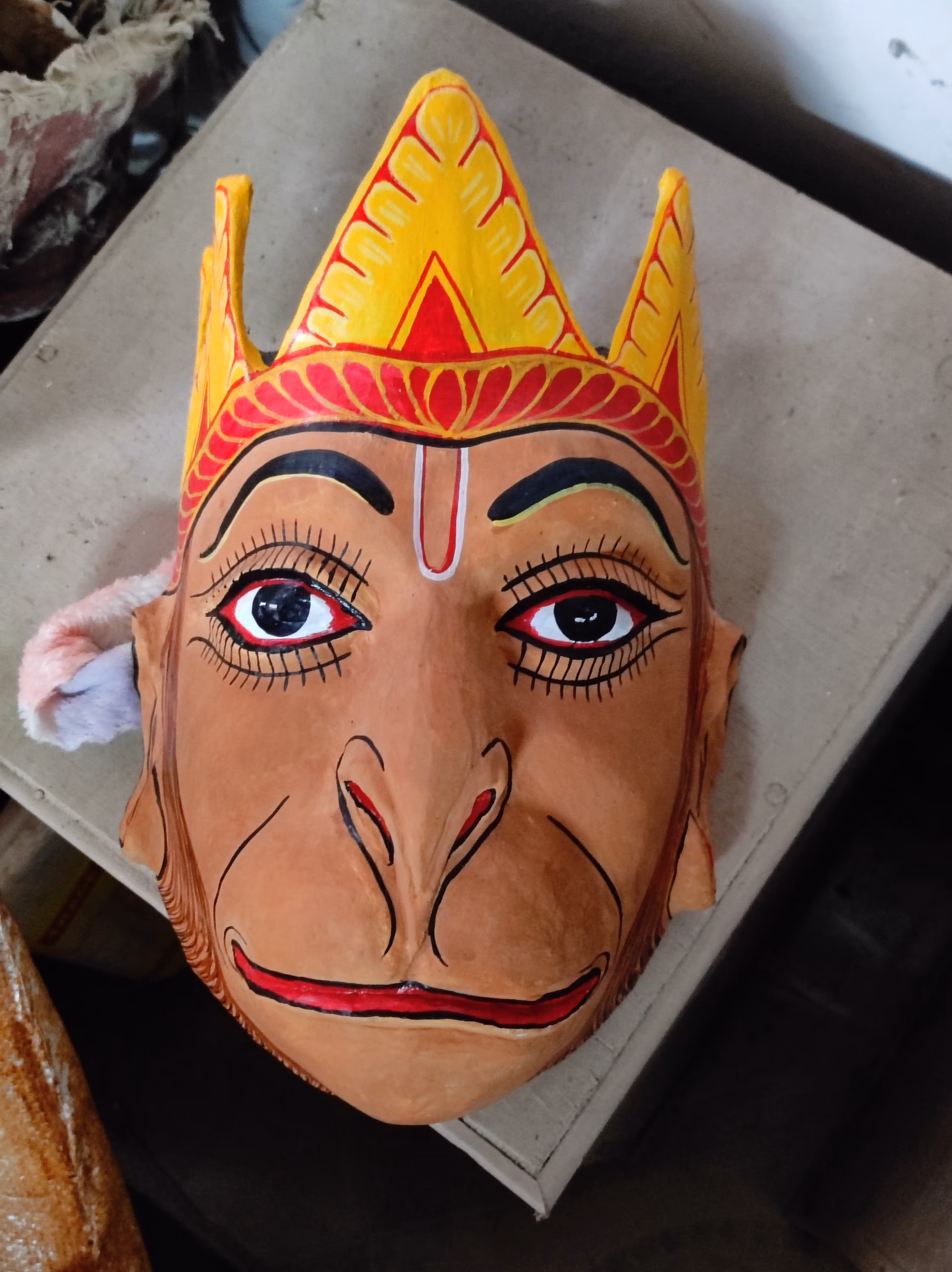 A monkey-faced Hanuman mukha resting on a plain surface, its long pink-and-ochre muzzle and wide painted eyes below a yellow-and-red pointed crown, a Majuli bhaona mask