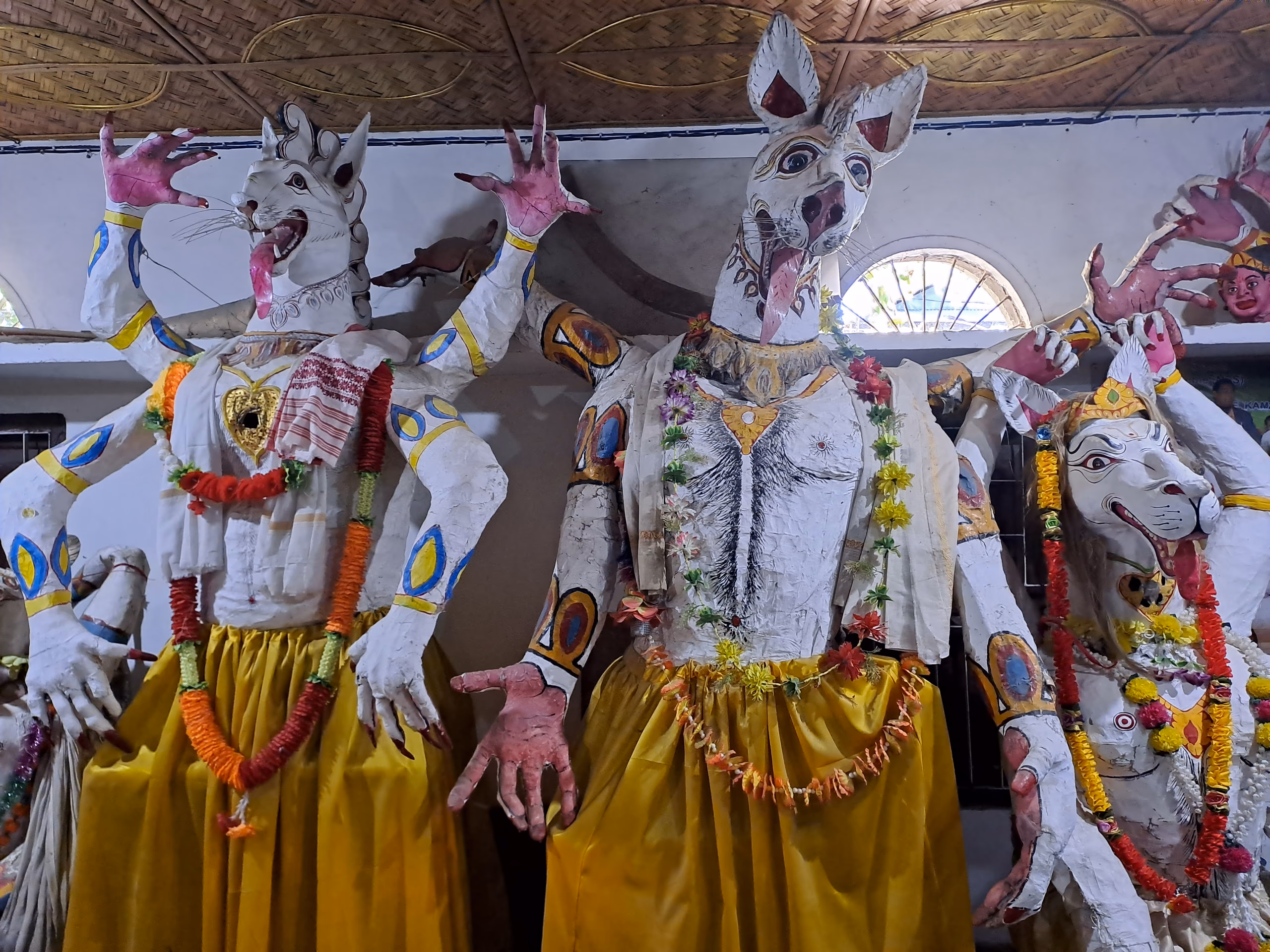 Three towering lion-headed cho-mukha body-masks at Samaguri Sattra, dressed in white jackets and yellow skirts, draped with marigold garlands and a gamosa, clawed hands raised, standing under the namghar's bamboo roof