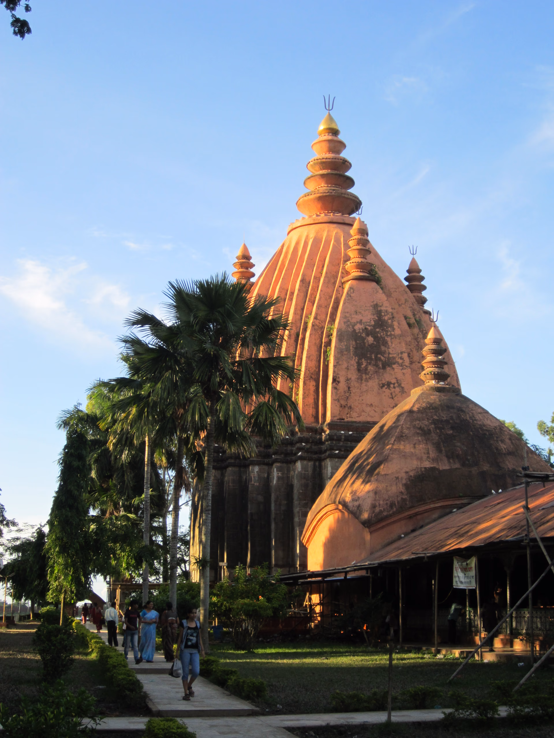 The towering red-ochre brick spire of the Sivadol at Sivasagar glowing in late-afternoon golden light against a deep blue sky, framed by a tall palm