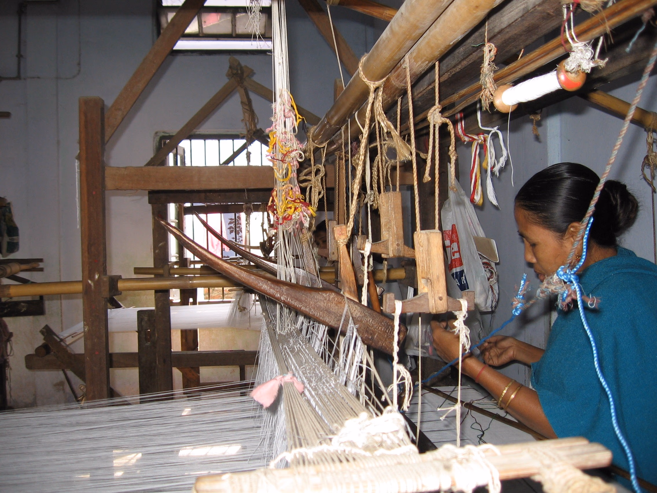 A woman weaving at a traditional wooden Assamese handloom, the warp threads fanning out beneath the heddles and shuttle batten as she works the cloth