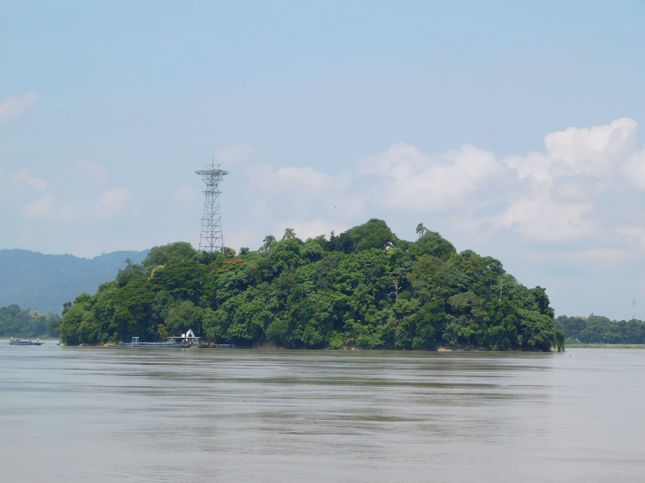 The wholly wooded Peacock Island rising from the broad Brahmaputra under a blue sky, the ferry landing at its near shore and low hills along the far bank