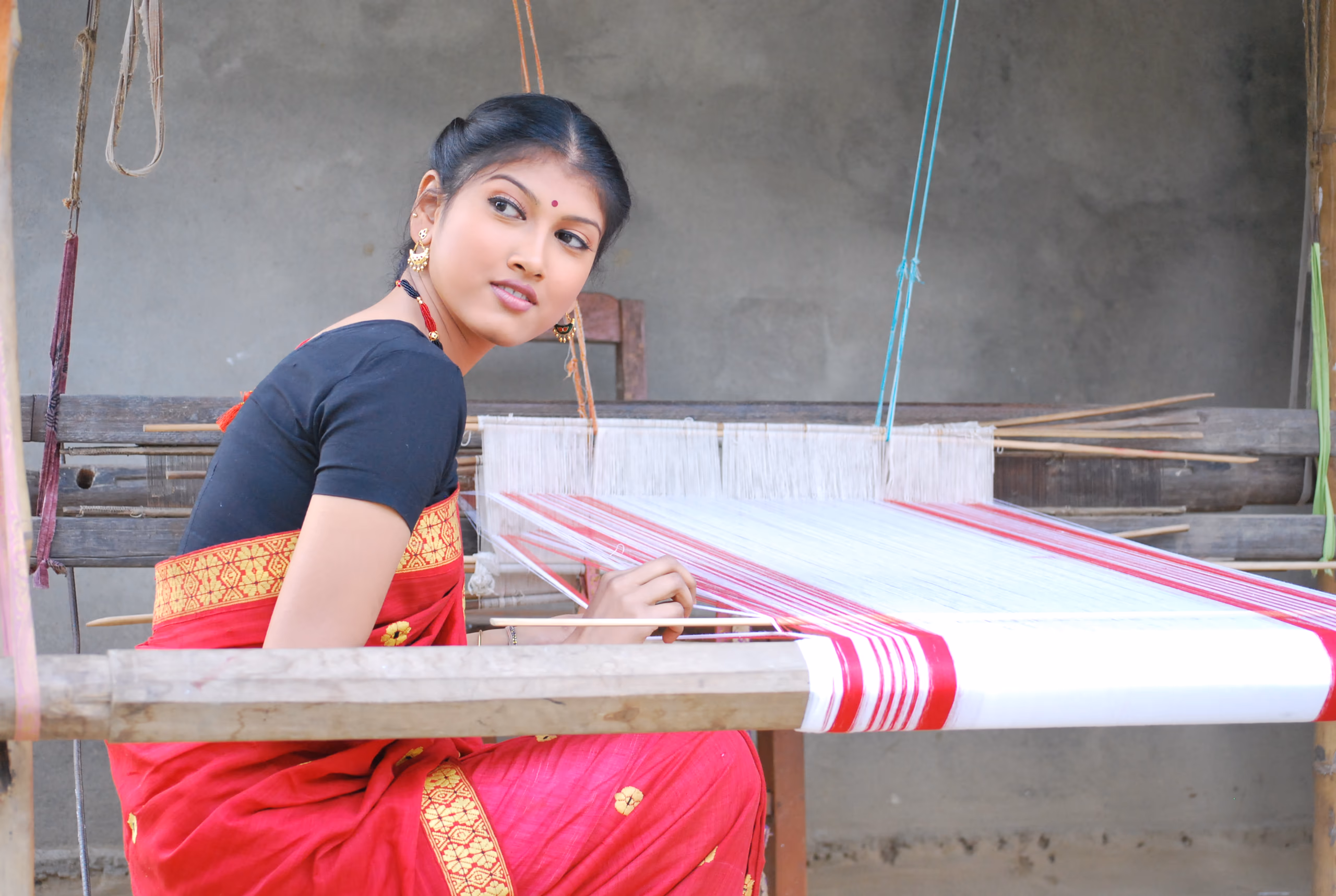 A woman in a traditional Assamese mekhela sador seated at a wooden handloom, a red-and-white gamosa taking shape in the warp before her